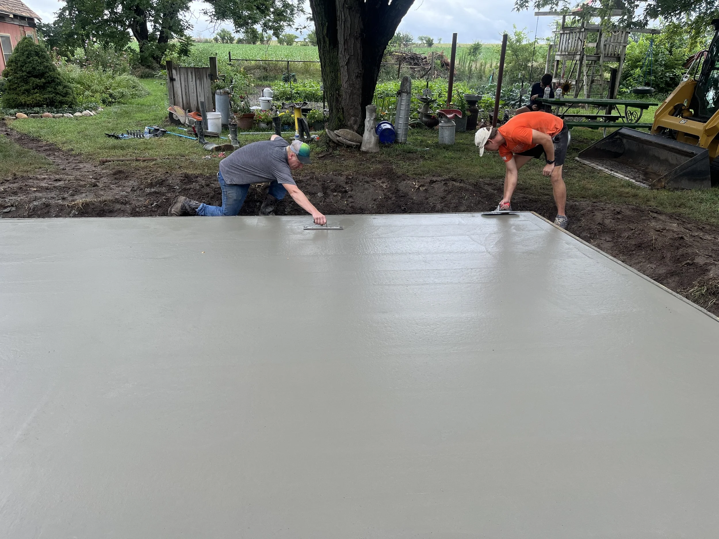 Two men are pouring and smoothing fresh concrete on a large outdoor surface in a backyard, with tools and gardening items scattered around.