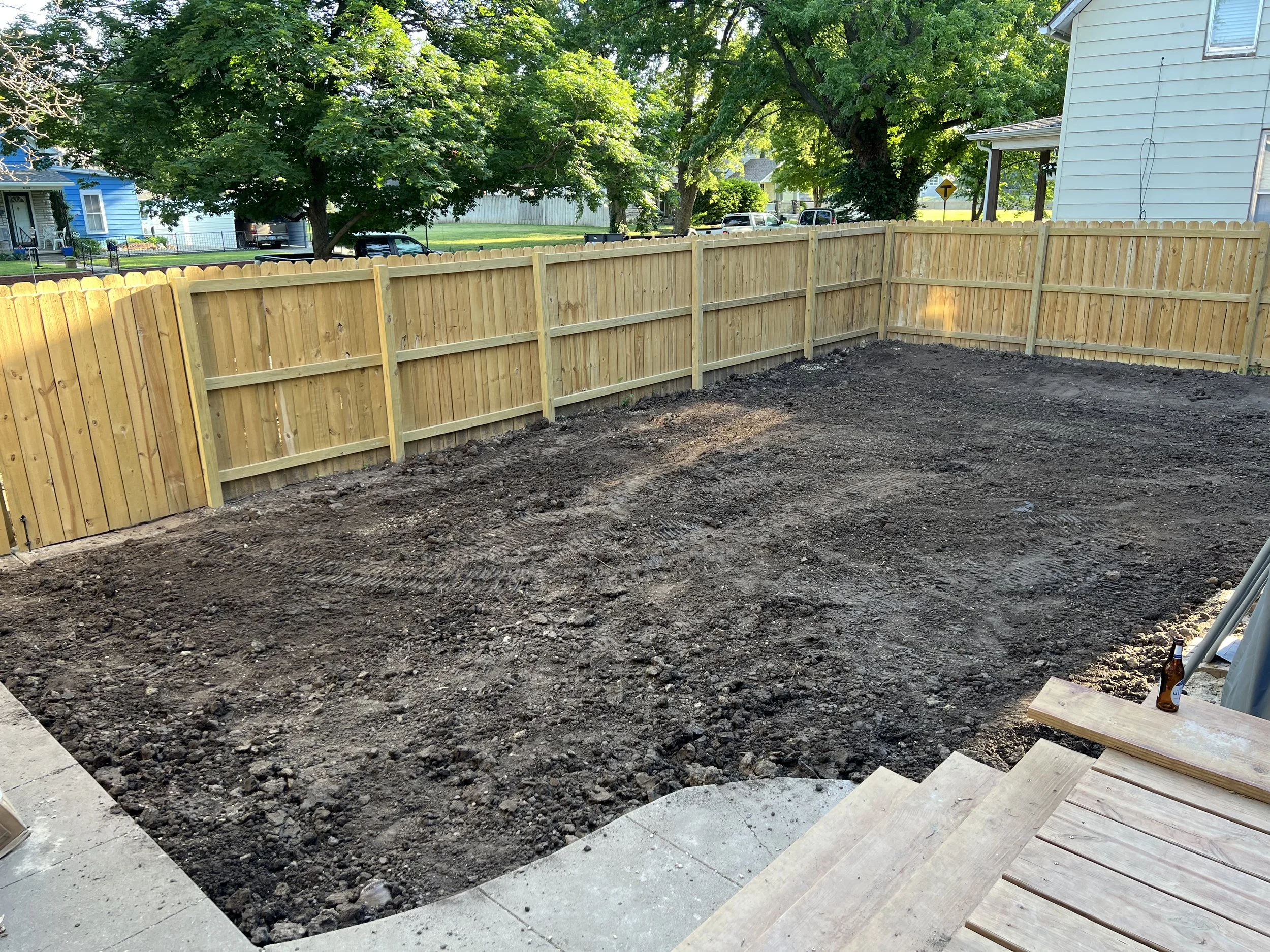 A backyard with a wooden privacy fence and a dirt ground, surrounded by trees and neighboring houses.
