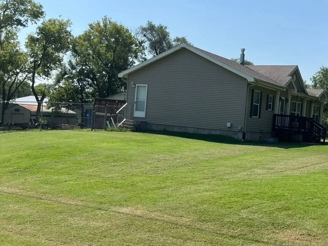 A house with beige siding, a small wooden deck, and a backyard with green grass and trees.