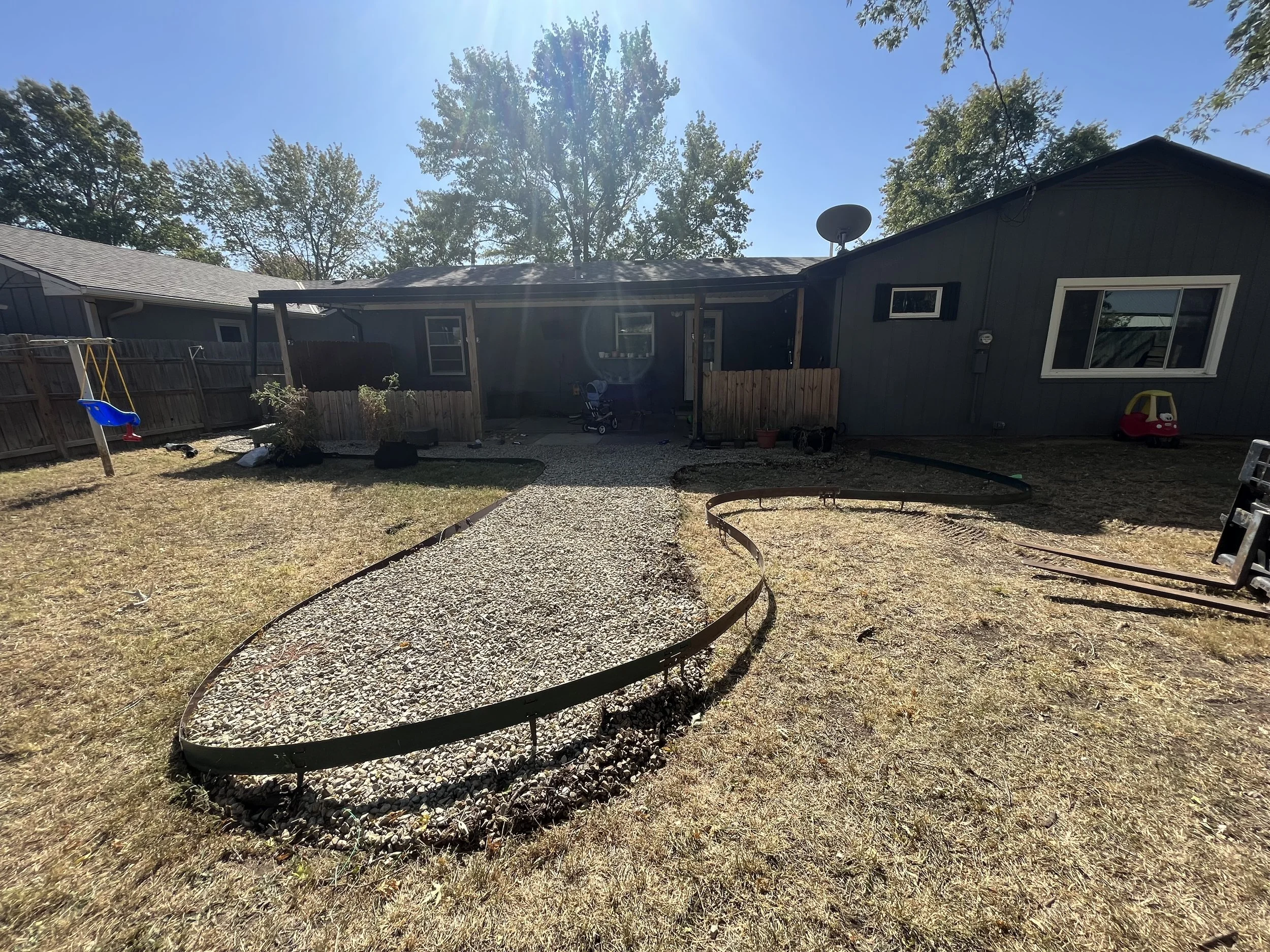 Backyard with gravel pathway, small play structures, and a covered patio. The house is dark-colored with white-framed windows, and tall trees can be seen in the background. The grass appears dry.