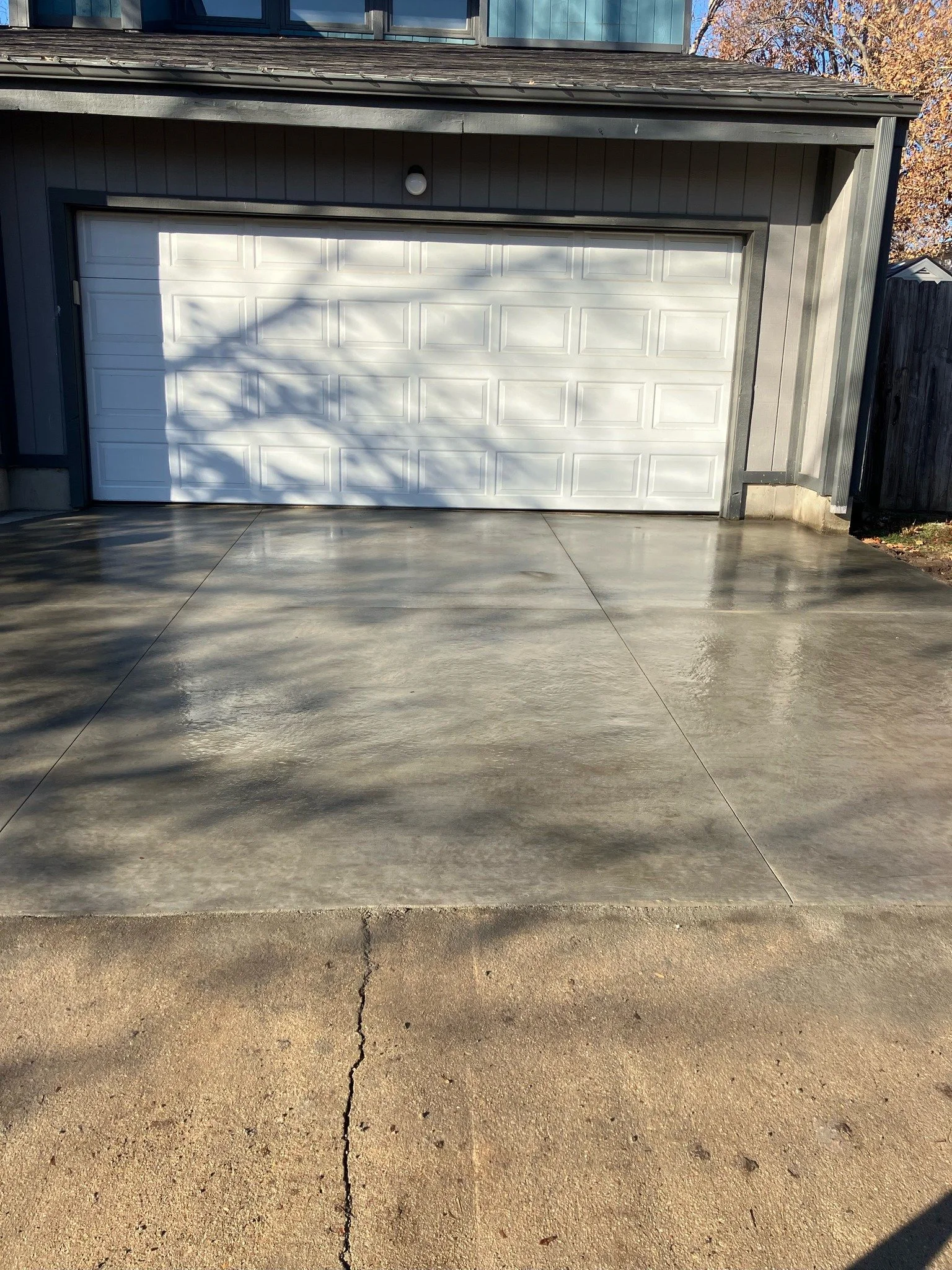 A clean, newly poured concrete driveway in front of a garage with a white door, gray siding, and a small outdoor light fixture. 