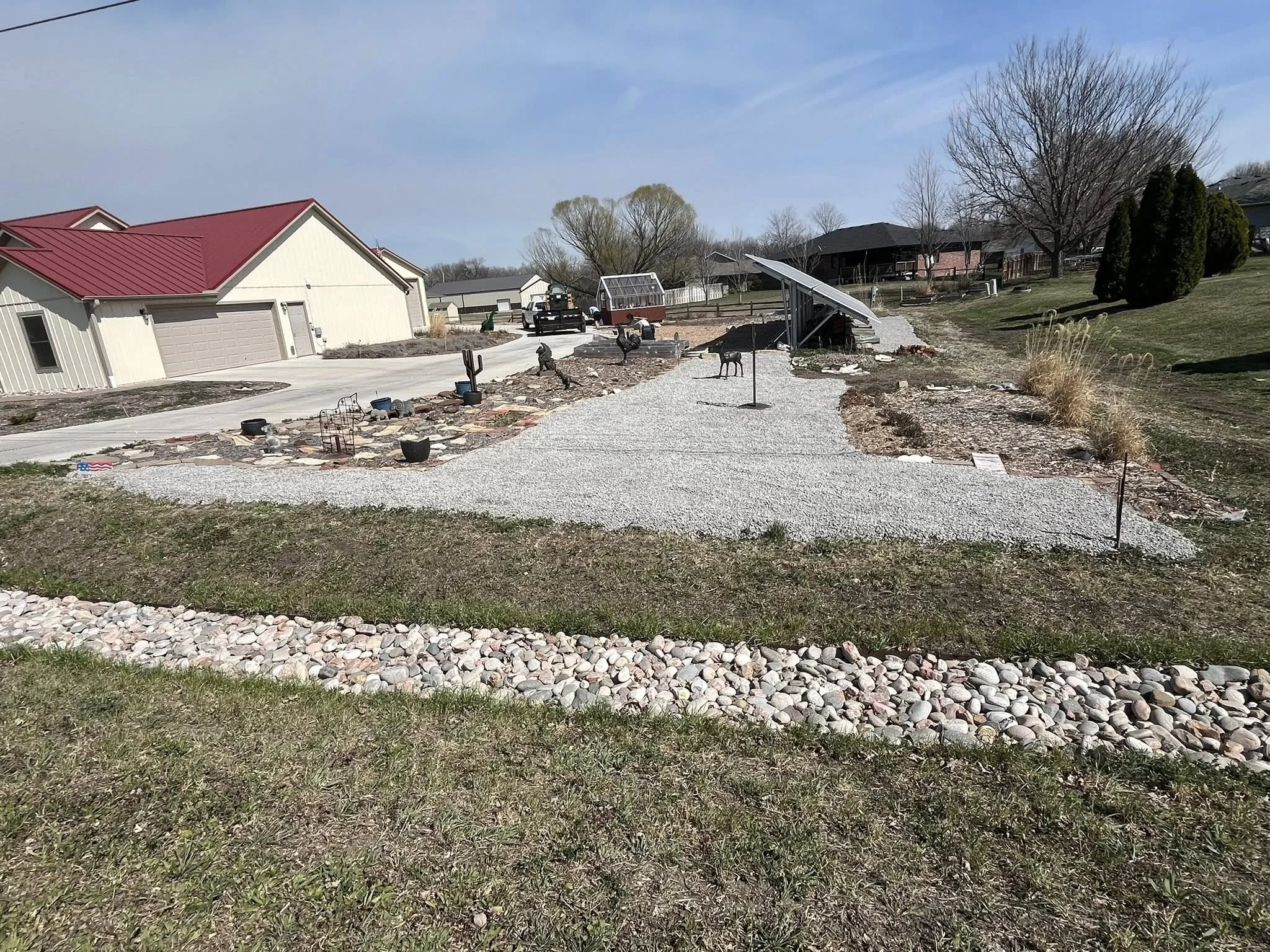 A backyard under construction with a gravel pathway, some rocks, and various garden decorations, with residential houses and trees in the background.