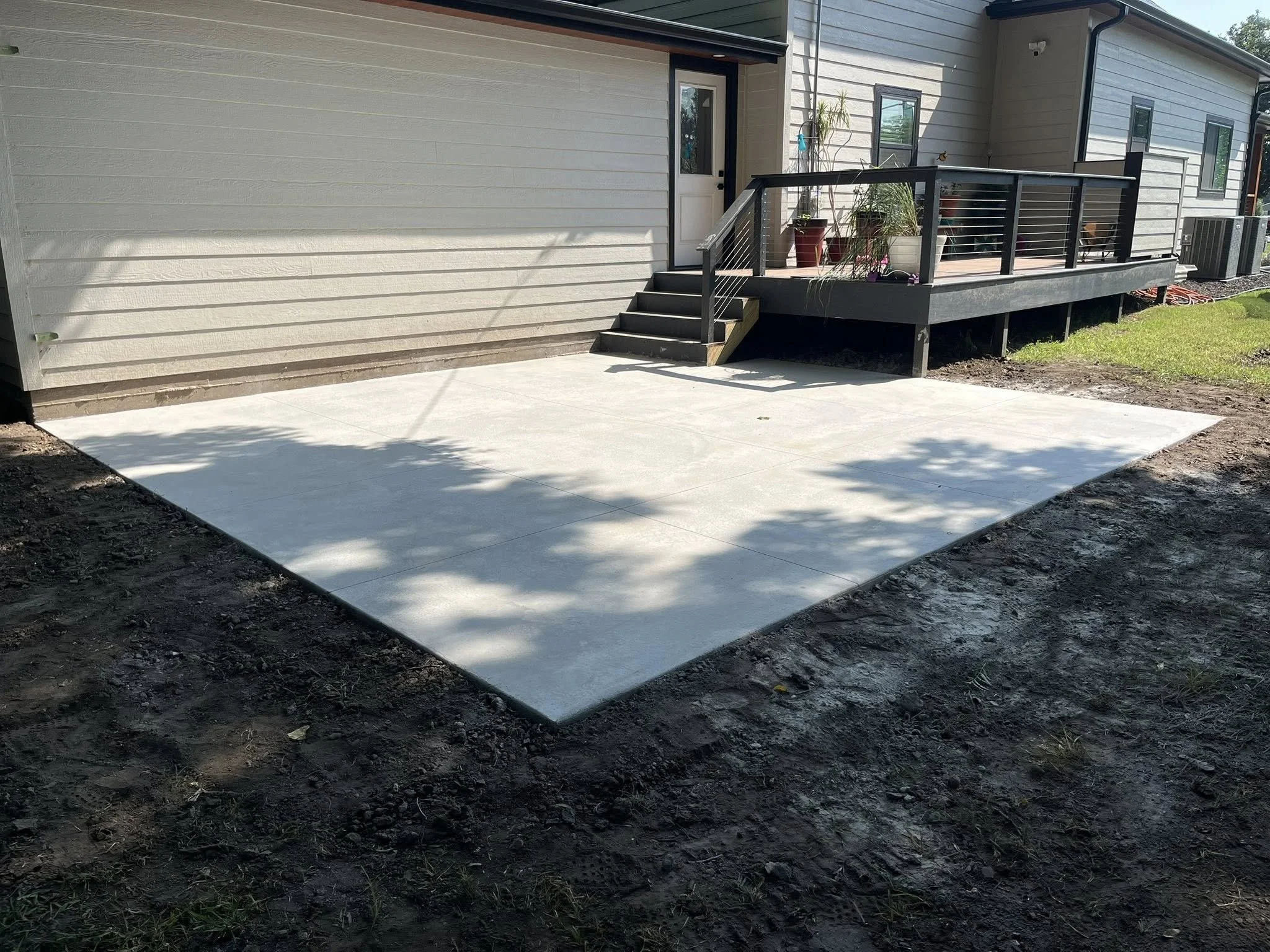 Concrete patio in backyard with a small staircase leading up to a deck attached to a house with beige siding and black railing, and potted plants on the deck.