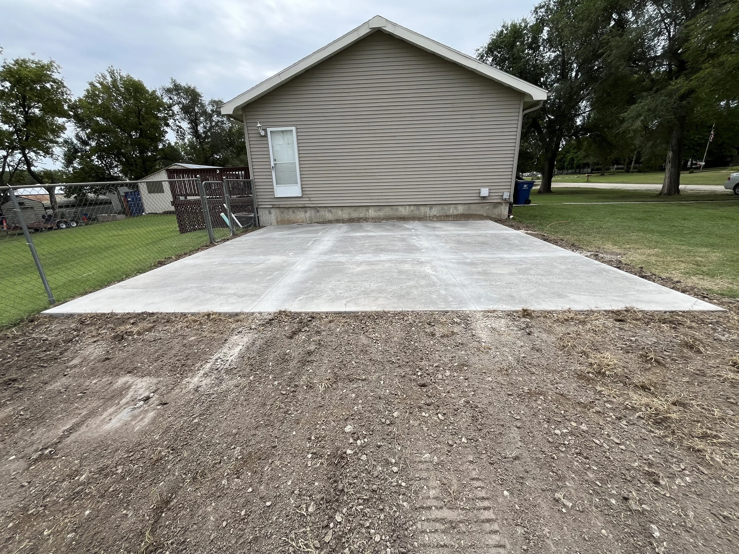 Concrete driveway in front of a beige house, fenced yard on the left, and grassy area with trees on the right.