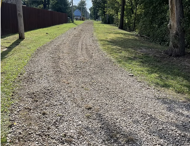A gravel path driveway running through a residential area with grass on both sides and trees on the right, with houses visible in the background.