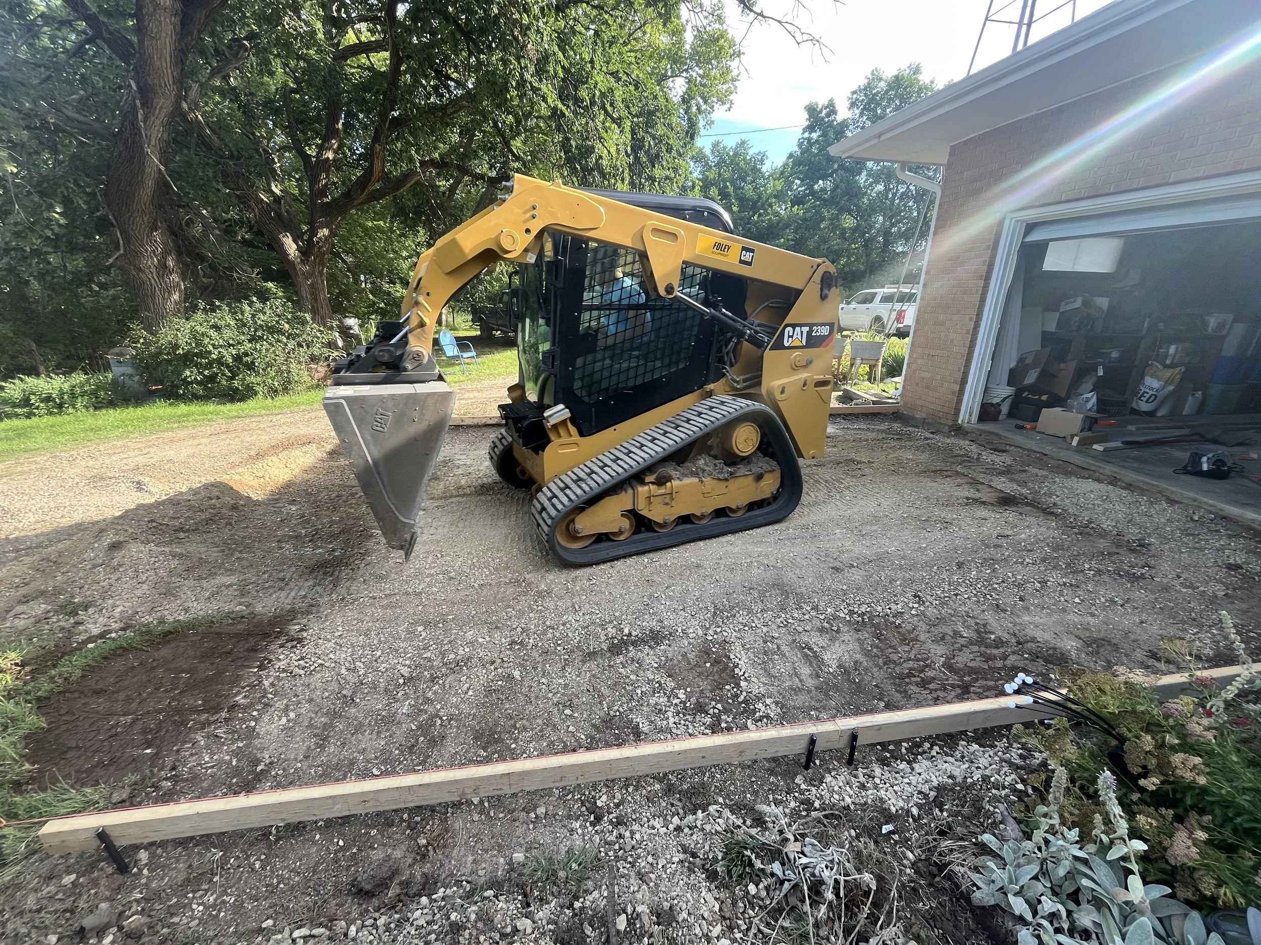 Small yellow Caterpillar CAT 239D compact track loader working on a gravel driveway outside a house with an open garage, trees in the background, and a rainbow streak in the sky.