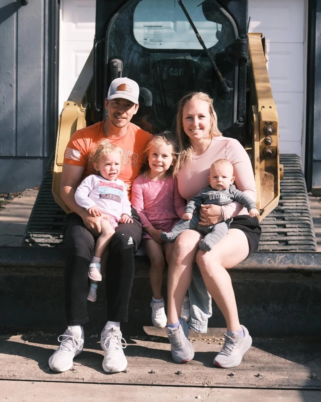 Family of five sitting in front of a construction vehicle, smiling at the camera. The family includes two adults and three children.