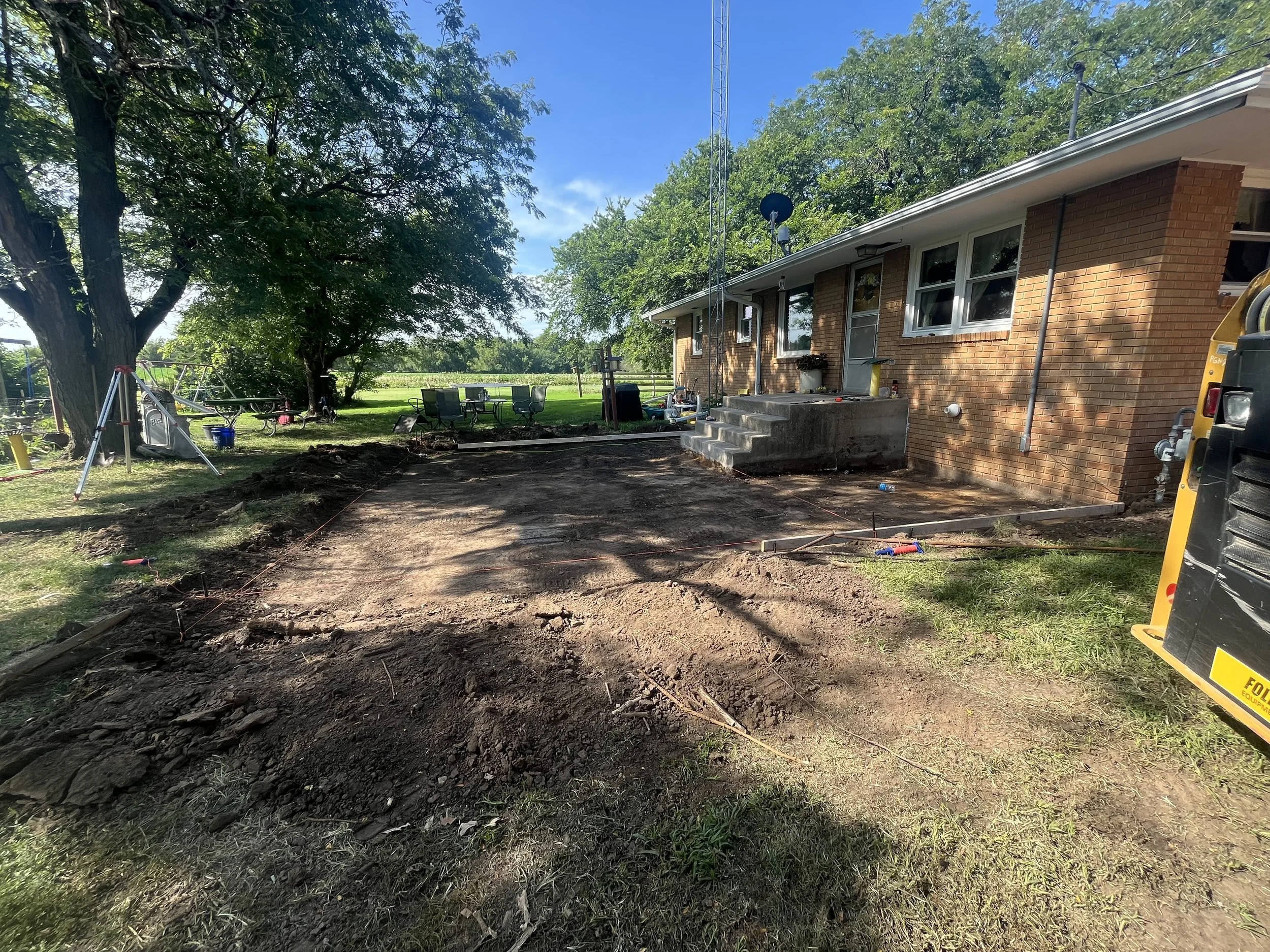 Backyard with freshly excavated dirt area and brick house with steps, surrounded by trees, outdoor furniture, and construction tools.