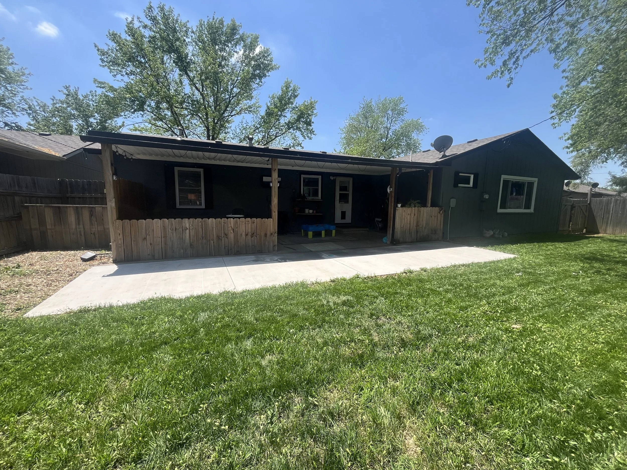 Backyard with a concrete patio, grass lawn, and a black house with a covered porch, surrounded by a wooden fence and tall trees.