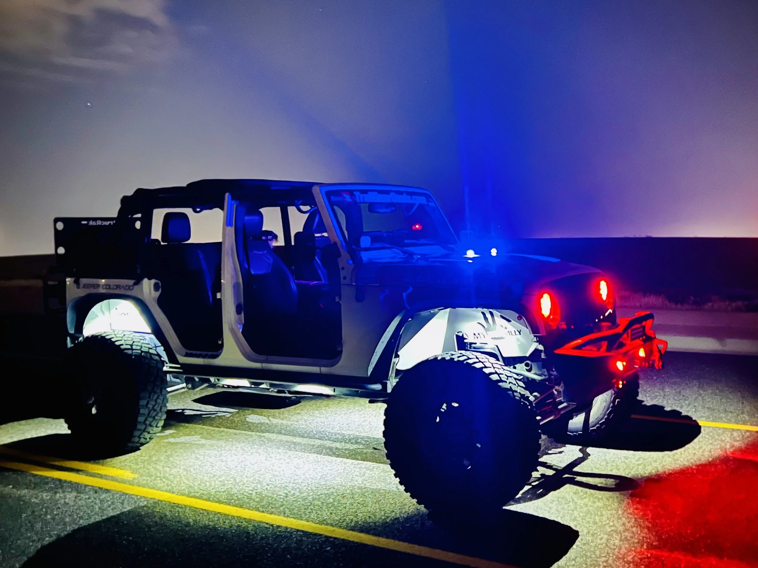 A dark-colored off-road vehicle, possibly a Jeep, with its doors open and emergency lights flashing, parked on a highway at night under a cloudy sky.