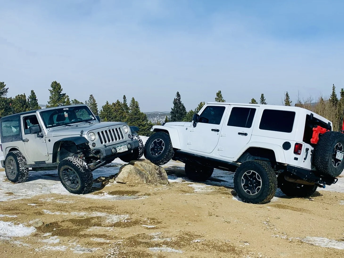 Two white Jeep vehicles on rocky, uneven terrain, with one vehicle balancing on a large rock, surrounded by trees and a partly cloudy sky.