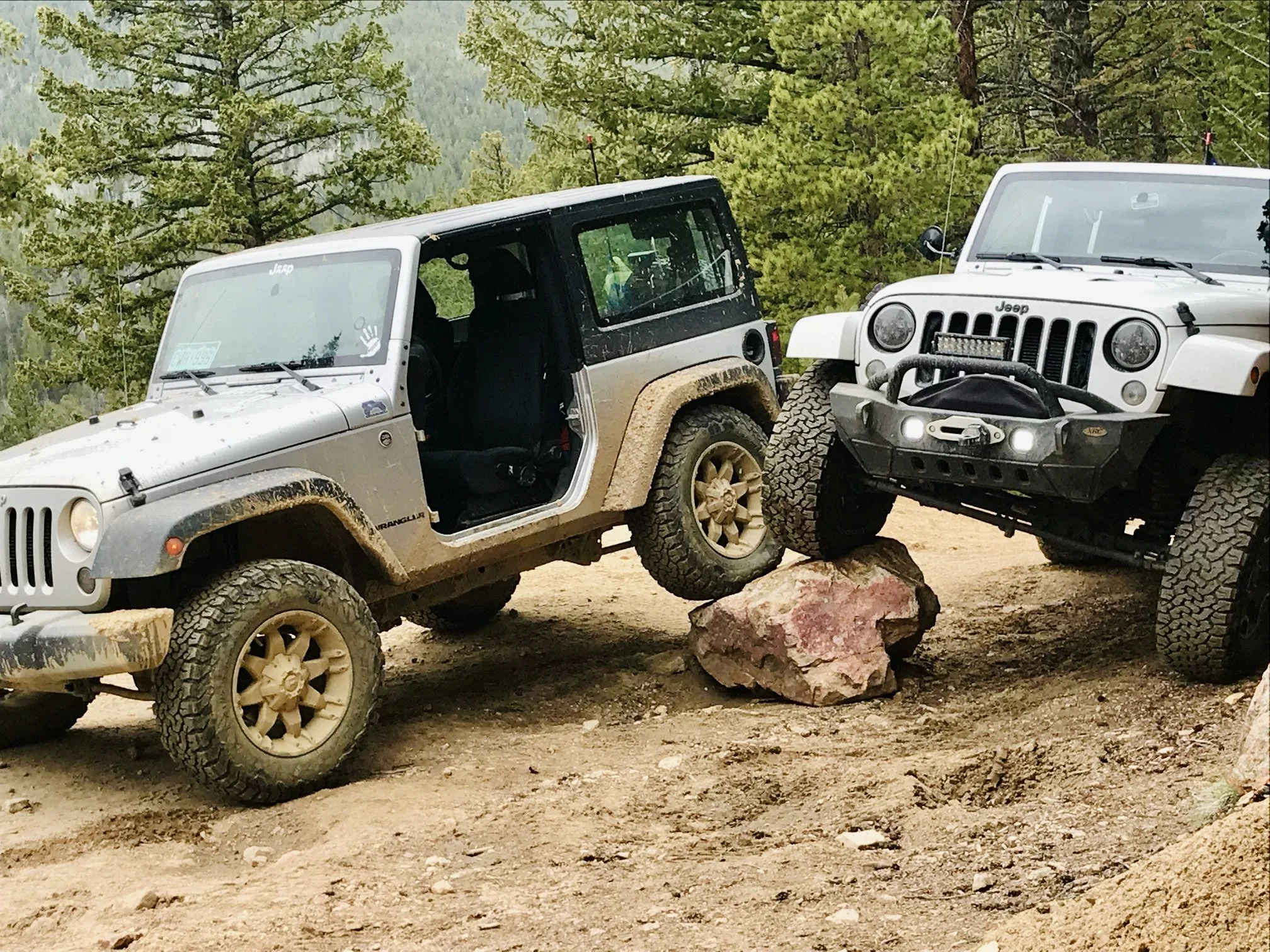 Two Jeep vehicles navigating off-road terrain in a forested area, with one Jeep's front wheels resting on a large rock, demonstrating rock crawling