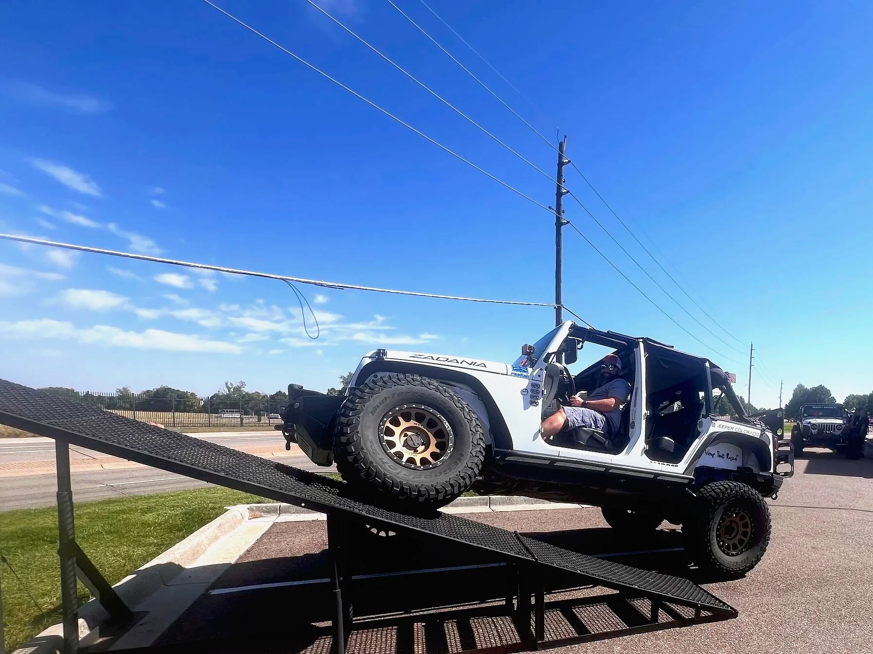 A white off-road vehicle on a ramp, with a driver wearing a helmet, preparing to descend a ramp during daytime with a blue sky and power lines overhead.