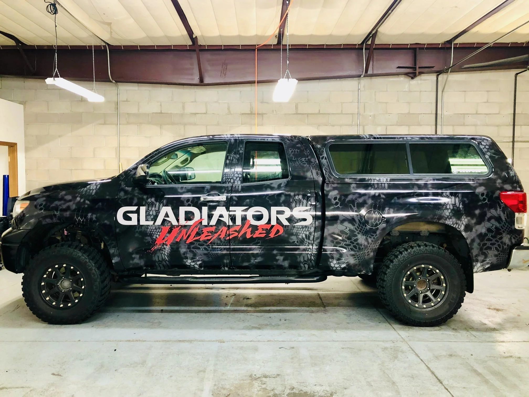 Black truck with leopard print pattern and large white and red text that says "Gladiators Unleashed" on the side, parked indoors against a brick and concrete wall.