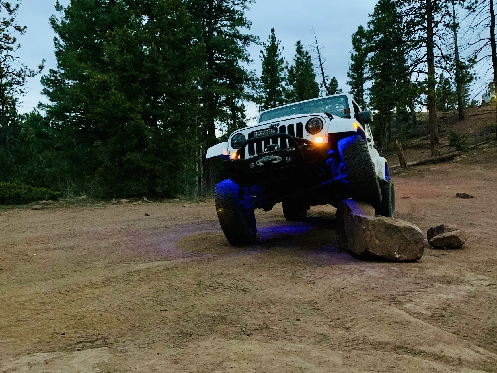 A white Jeep off-road vehicle with large tires balancing on a large rock in a forested area during twilight.