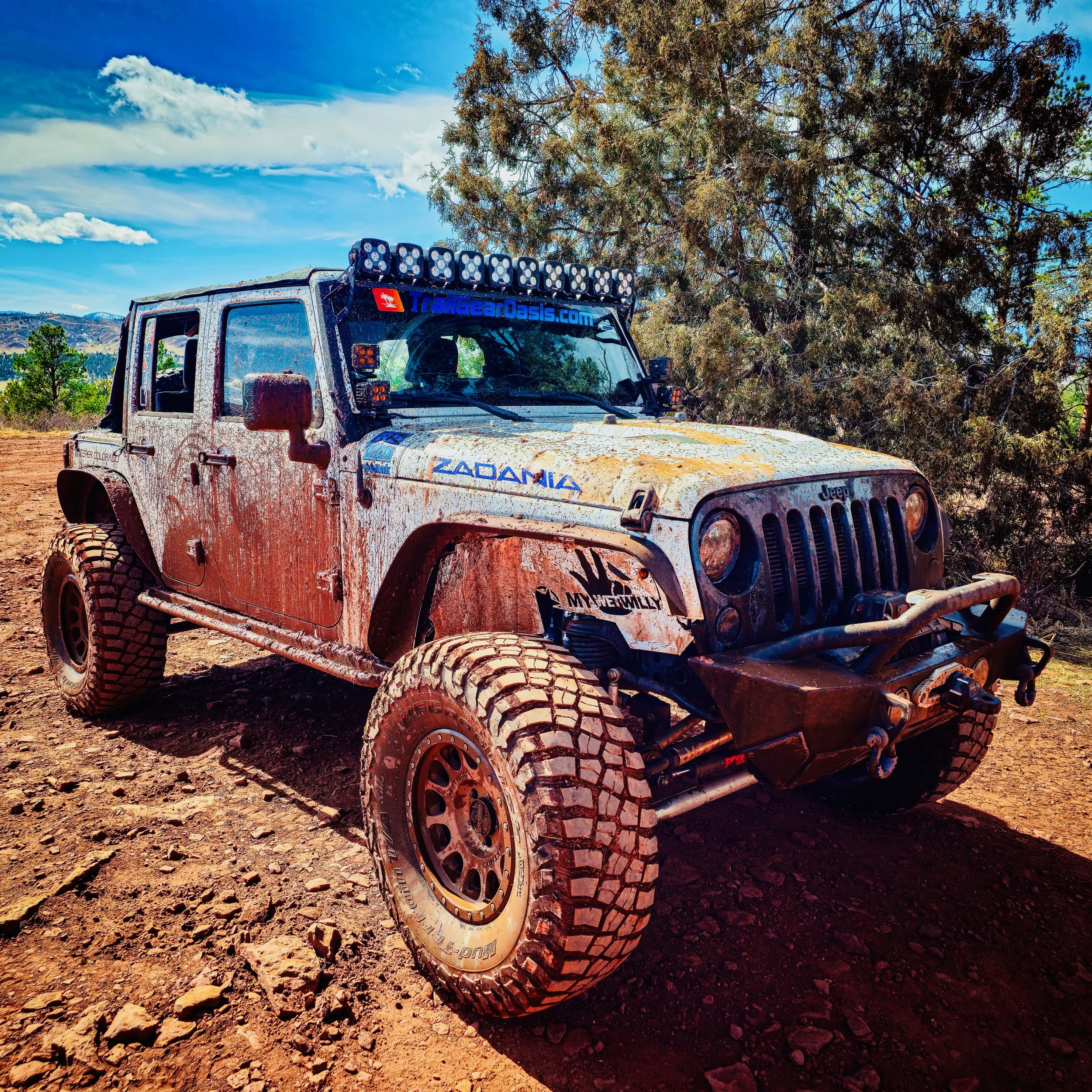 A muddy off-road Jeep in a dirt trail with trees and blue sky in the background.