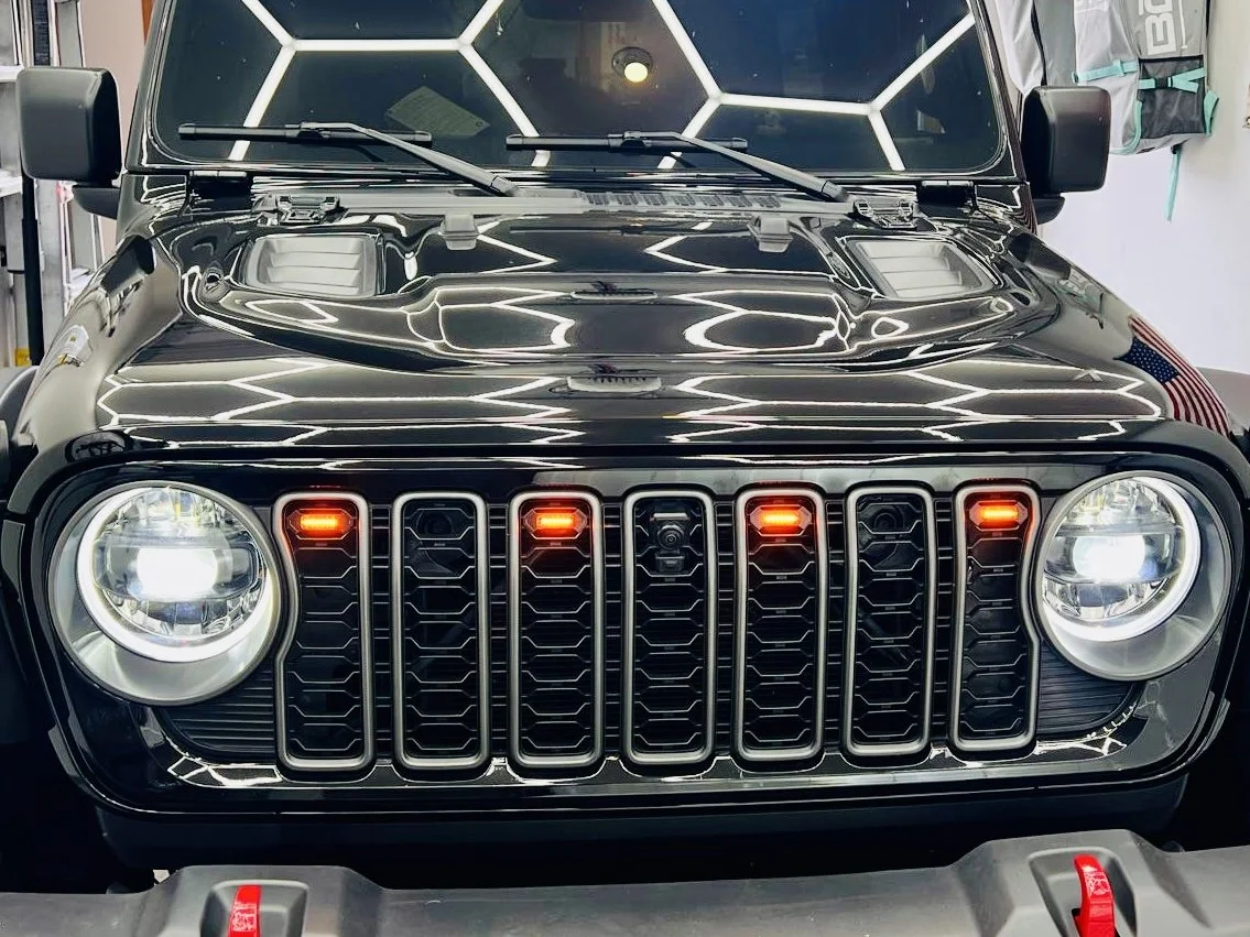 Front view of a black Jeep with illuminated headlights and seven-slot grille, featuring red indicator lights on the grille and a partial view of a backpack on the side.