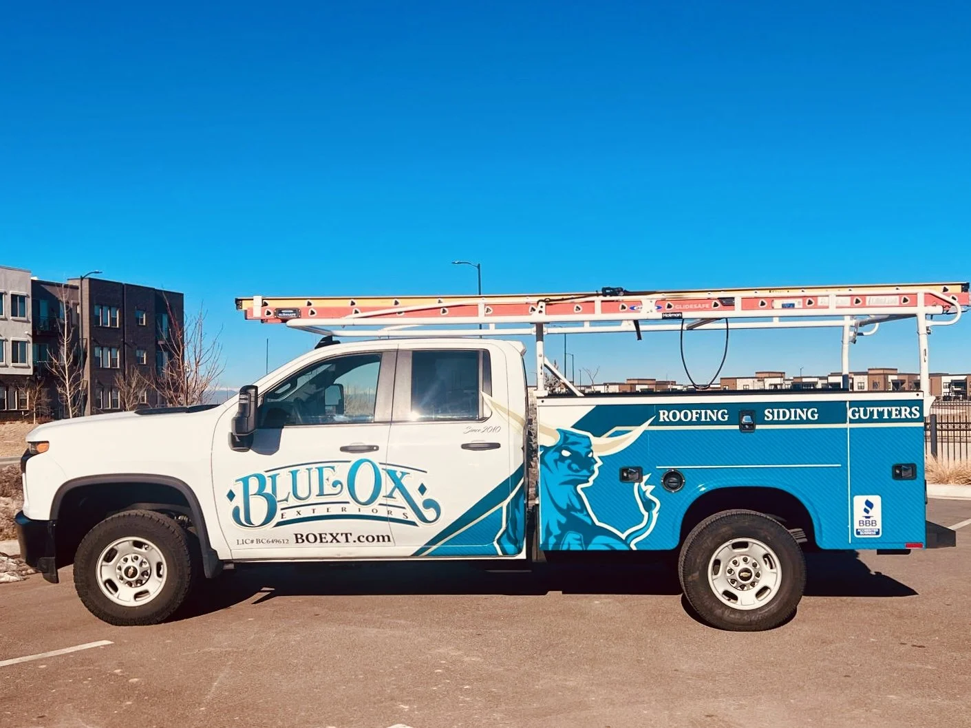 Service truck for Blue Ox Exteriors with a picture of a blue ox and logo. The truck has logos indicating roofing, siding, and gutter services, along with a ladder on top.