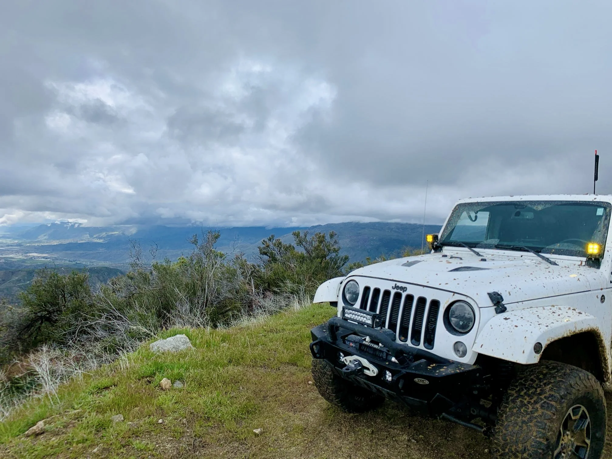 White Jeep off-road vehicle on grassy hillside with mountains and cloudy sky in the background.