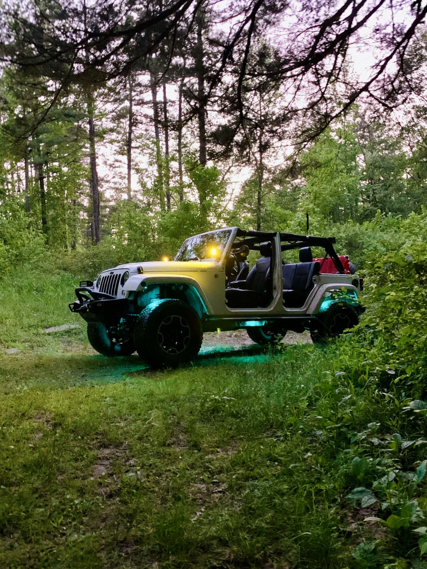 A white off-road Jeep with open doors parked in a forest clearing at dusk, illuminated by colored underglow lights