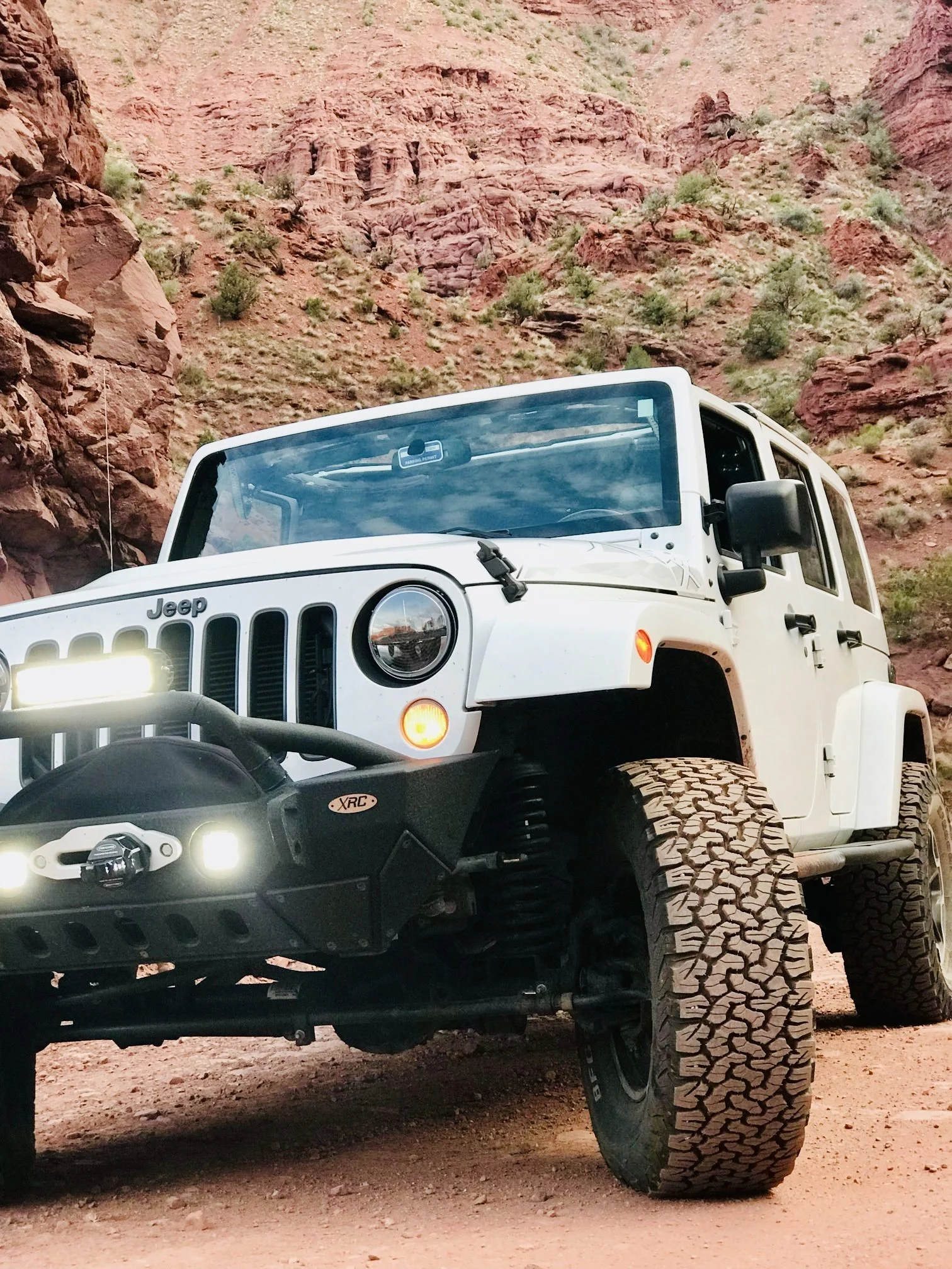 A white Jeep Wrangler off-road vehicle with large tires parked on a dirt trail against a backdrop of red rocky cliffs and sparse green vegetation.