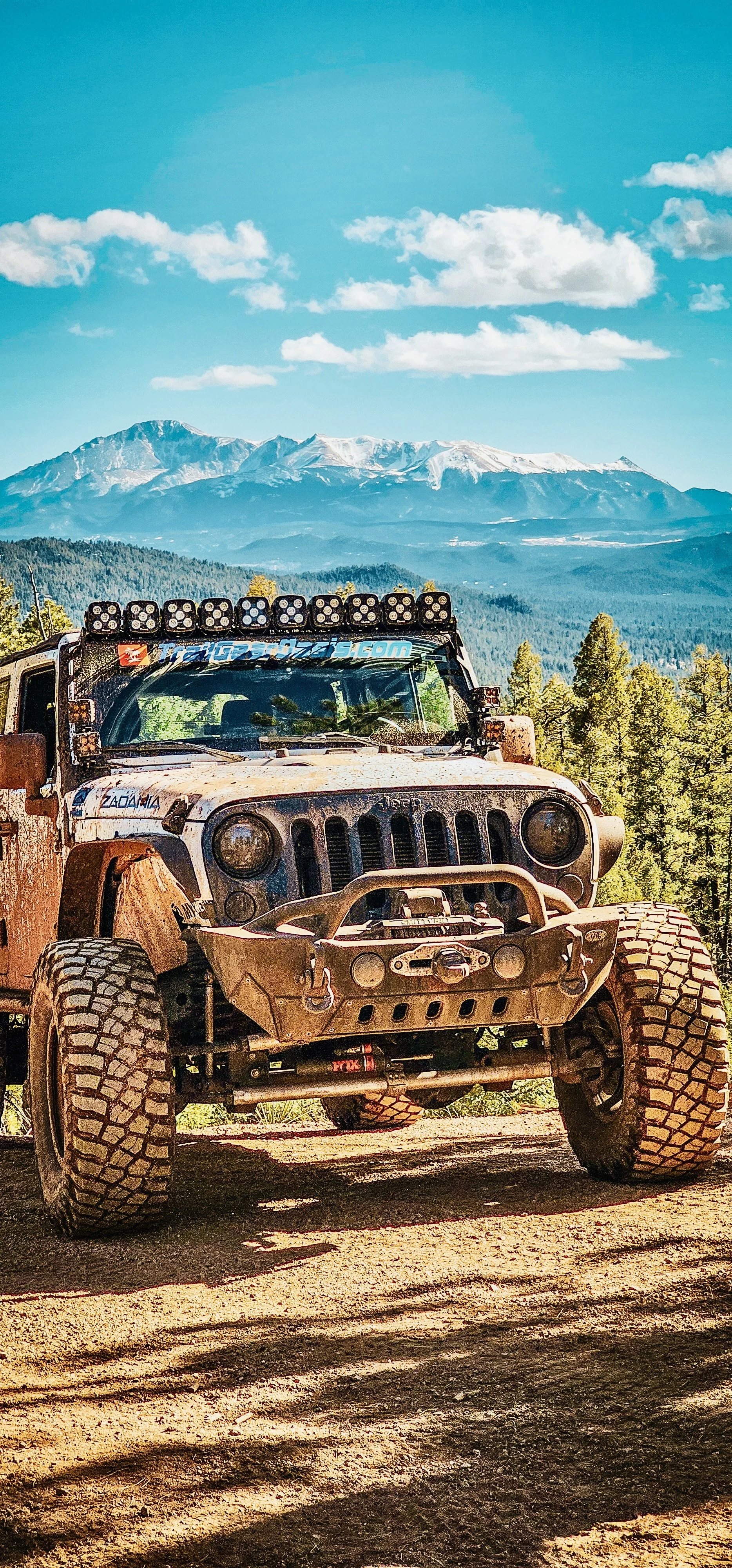 A rugged off-road Jeep vehicle with dirt on its body, large tires, and roof-mounted lights, parked on a dirt trail in a mountainous forest area with snow-capped peaks and blue sky in the background.