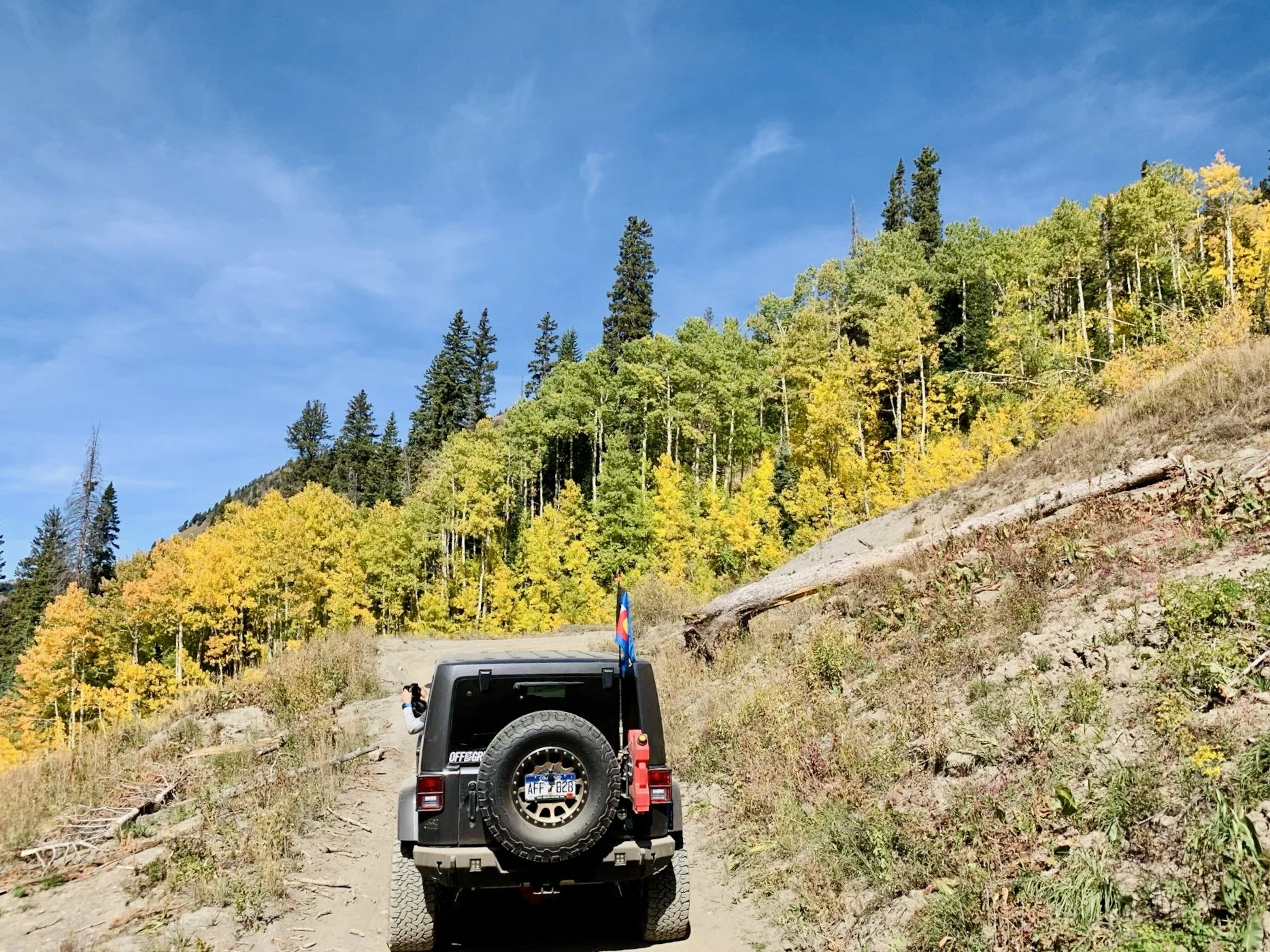 A black off-road vehicle on a dirt trail in a mountainous forest with green and yellow trees, under a blue sky.