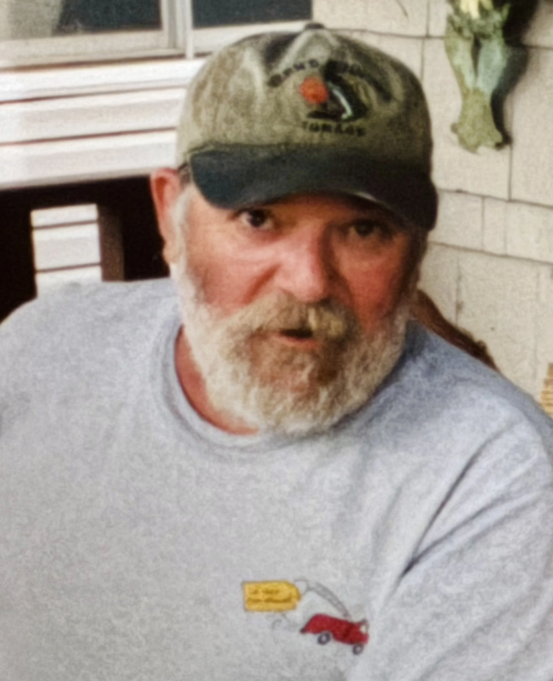 A bearded man wearing a gray shirt with a small yellow and red logo on it, a baseball cap with an image of a turkey on the front, sitting in front of a brick wall and window.