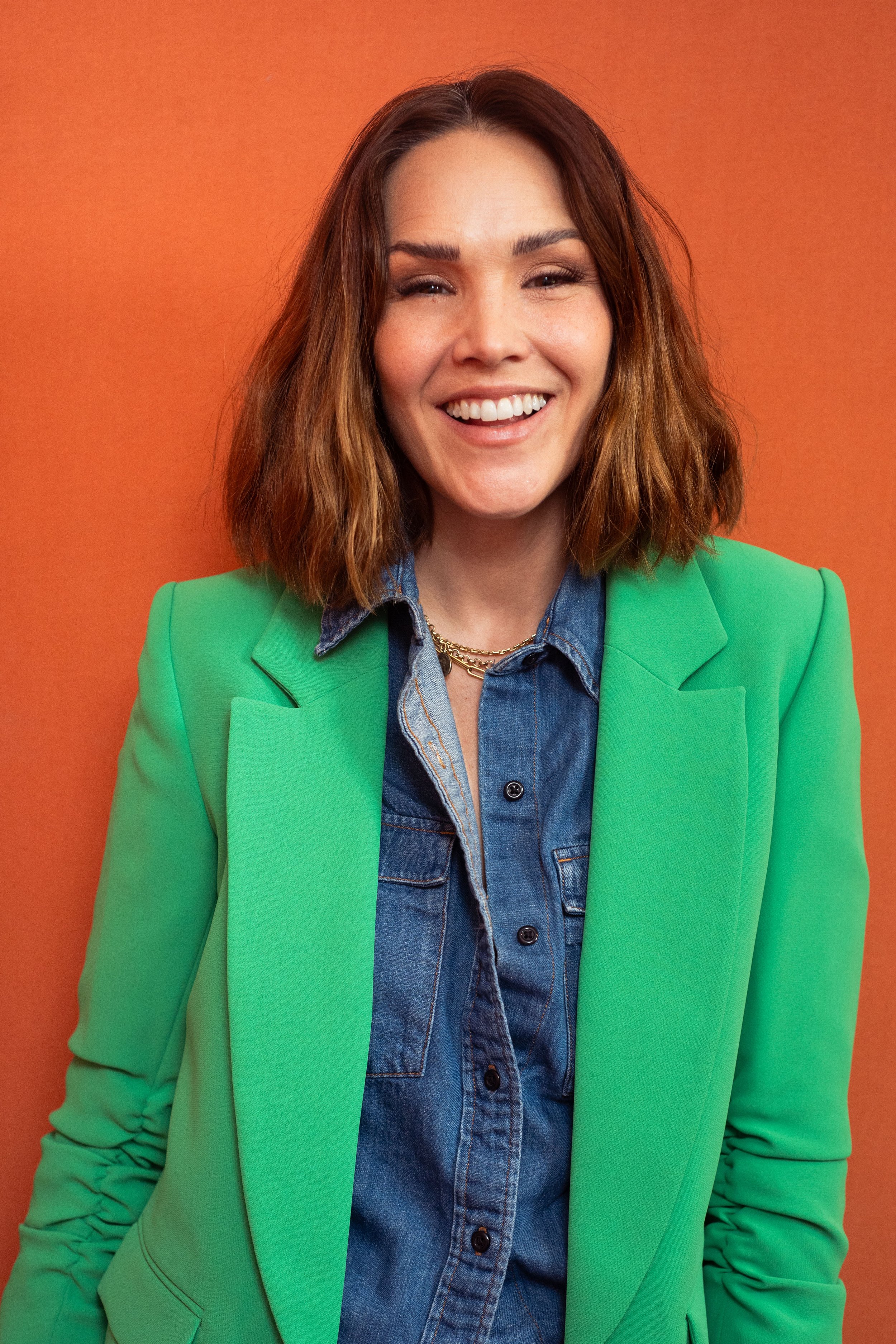 Mujer sonriendo con cabello castaño y rizado, usando saco verde y camisa de mezclilla, frente a fondo anaranjado.
