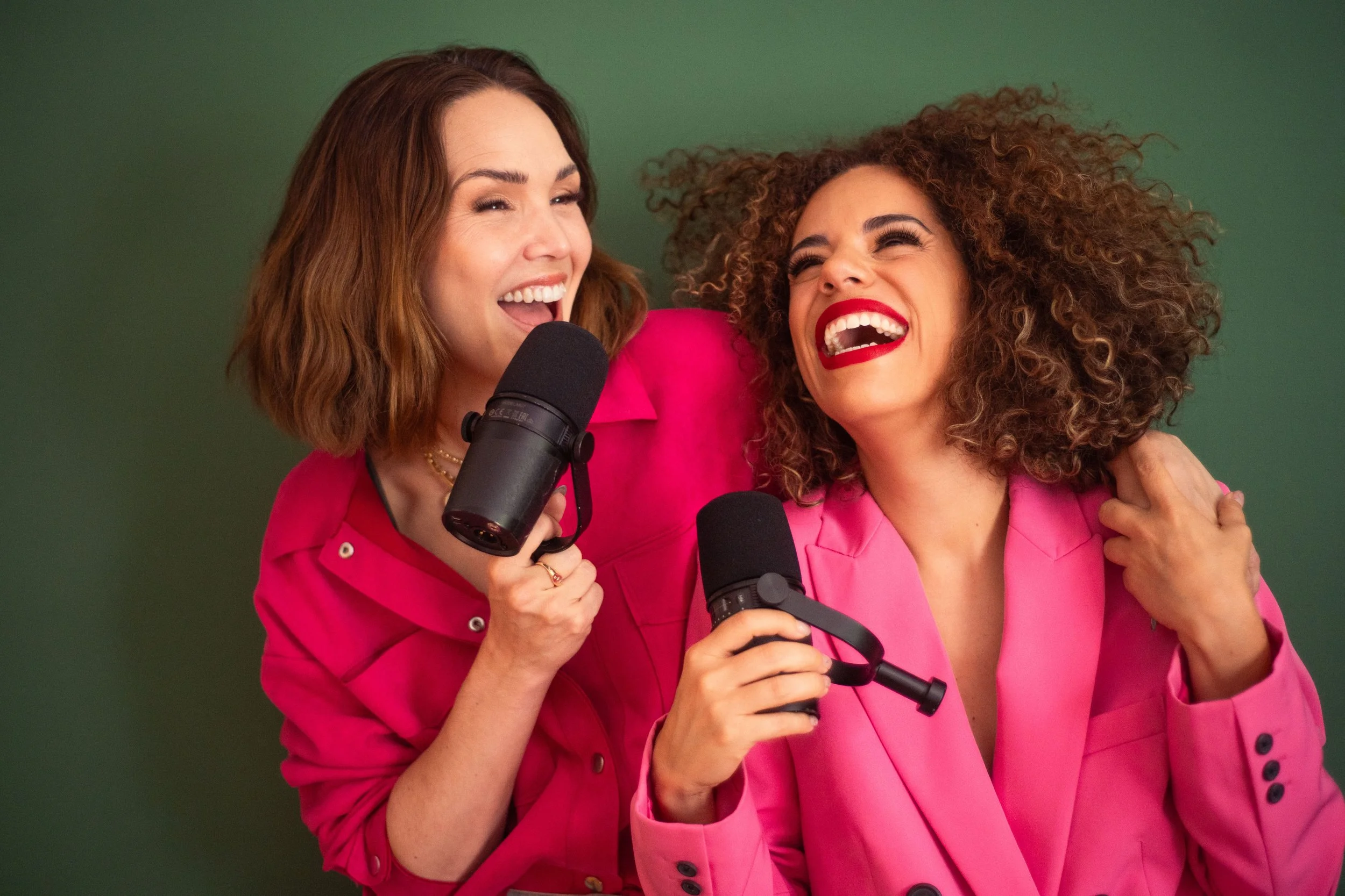 Dos mujeres jóvenes riendo, una con cabello castaño y la otra con cabello rizado, ambas con ropa rosa, sosteniendo micrófonos.