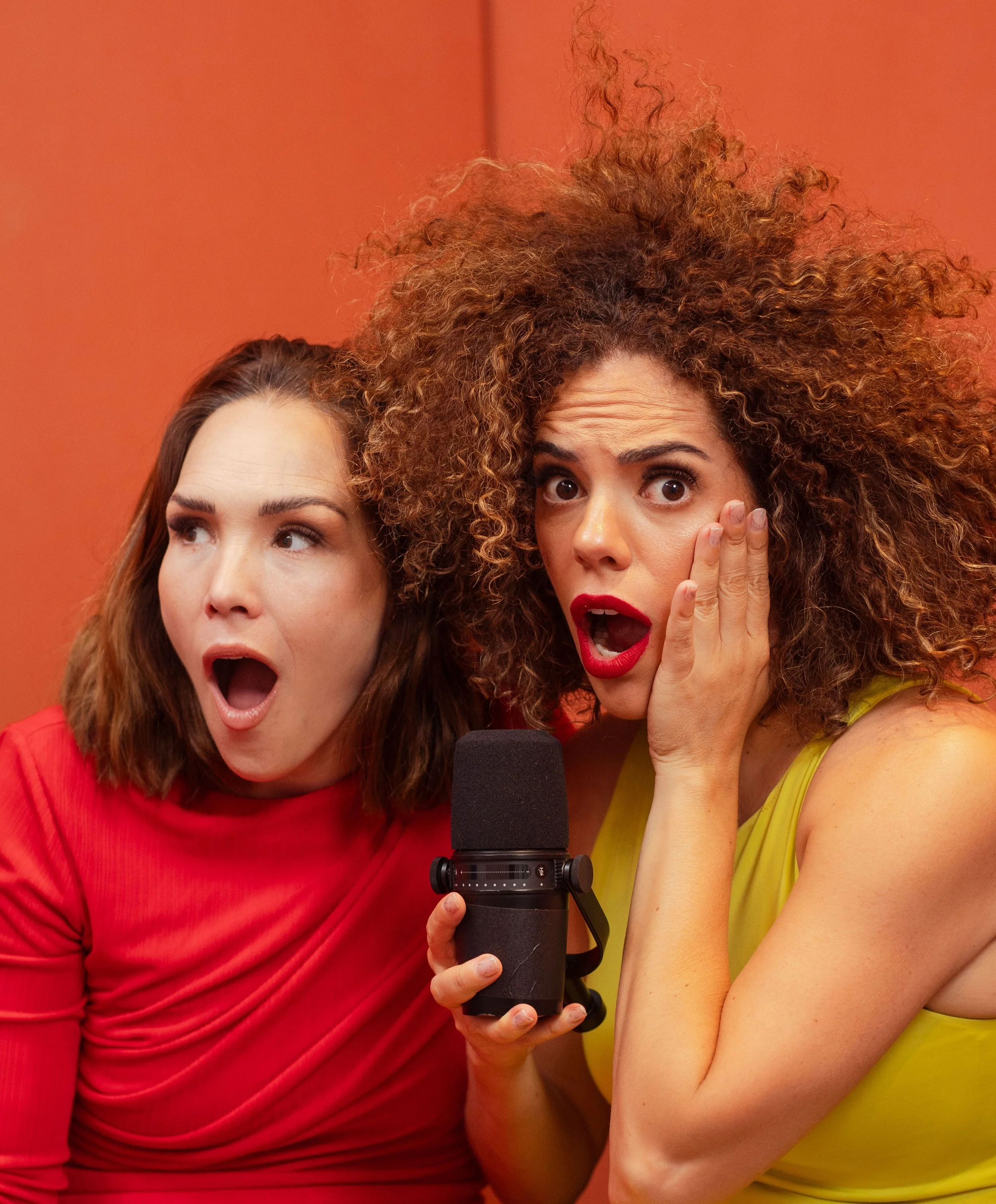 Dos mujeres (Priscila Faz y Alexandra Ivanisevic, hosts de Sepa la madre Podcast) con expresiones de sorpresa, una con vestido rojo y otra con vestido amarillo, sosteniendo un micrófono en un fondo naranja.