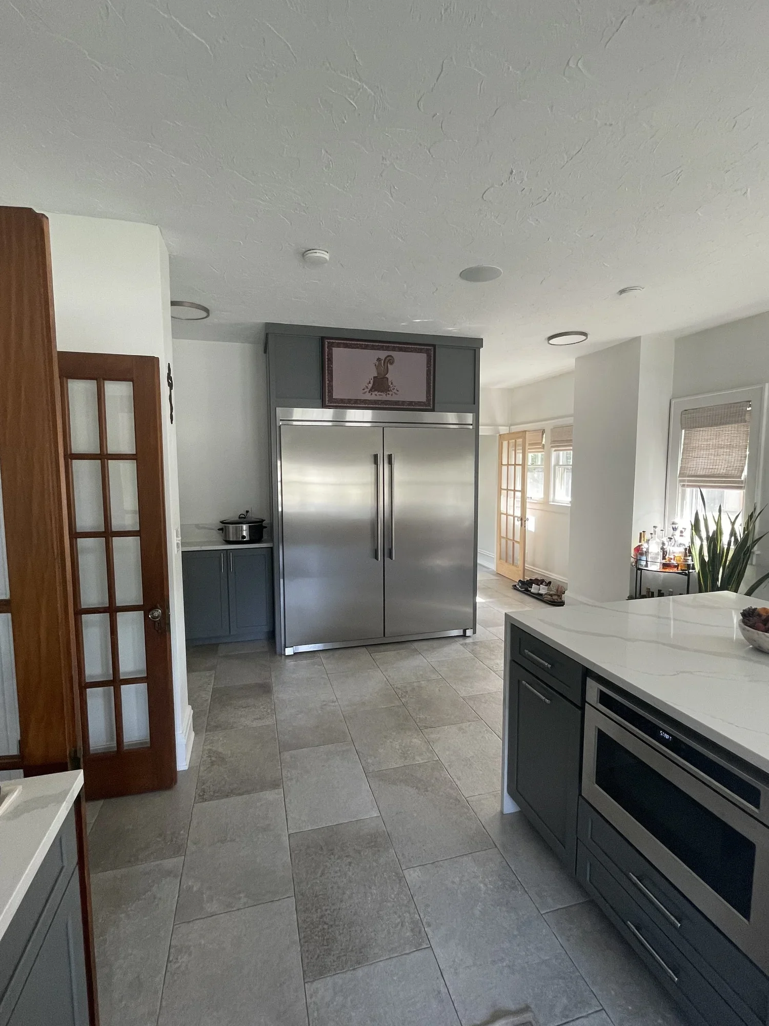 Modern kitchen remodel featuring a built-in stainless steel refrigerator surrounded by gray shaker cabinetry, white quartz countertops, gray porcelain tile flooring, a microwave drawer in the island, French doors, and abundant natural light.
