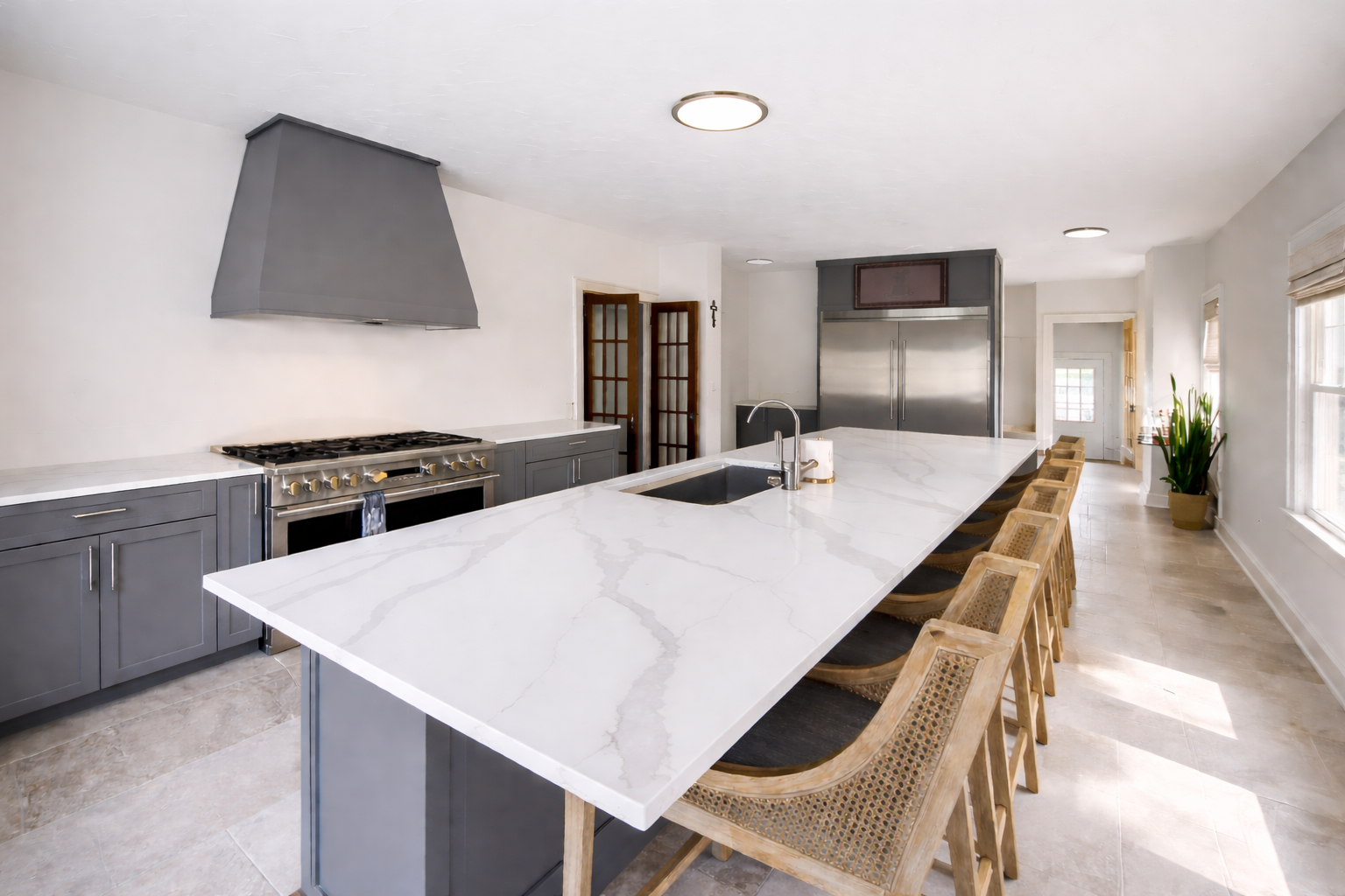 photo shows a large kitchen countertop island in a bright and airy kitchen -- there are cane and wood chairs lining the counter and a contrasting grey oven vent hood