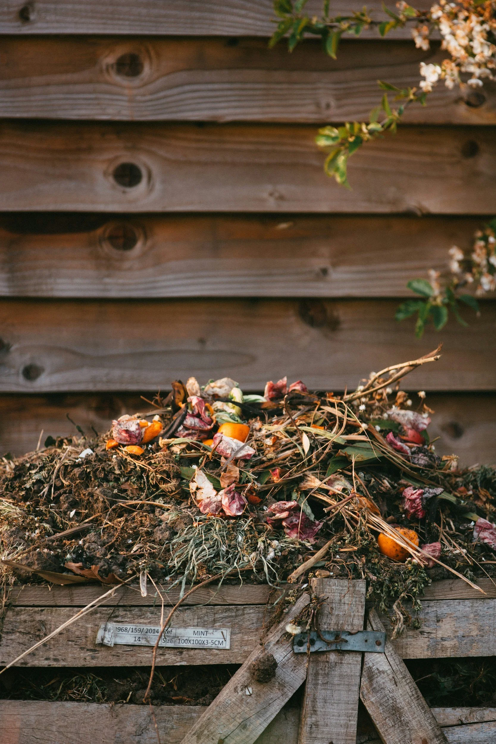 A pile of compost with organic waste such as vegetable scraps, leaves, and twigs in front of a wooden compost bin.