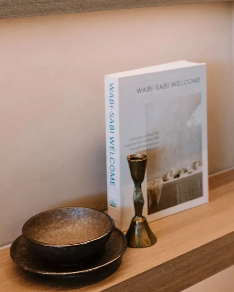 A wooden shelf holding a stack of three ceramic bowls, a black and gold chalice, and a white book with blue text titled 'WABI-SABI WELCOME' leaning against a beige wall.