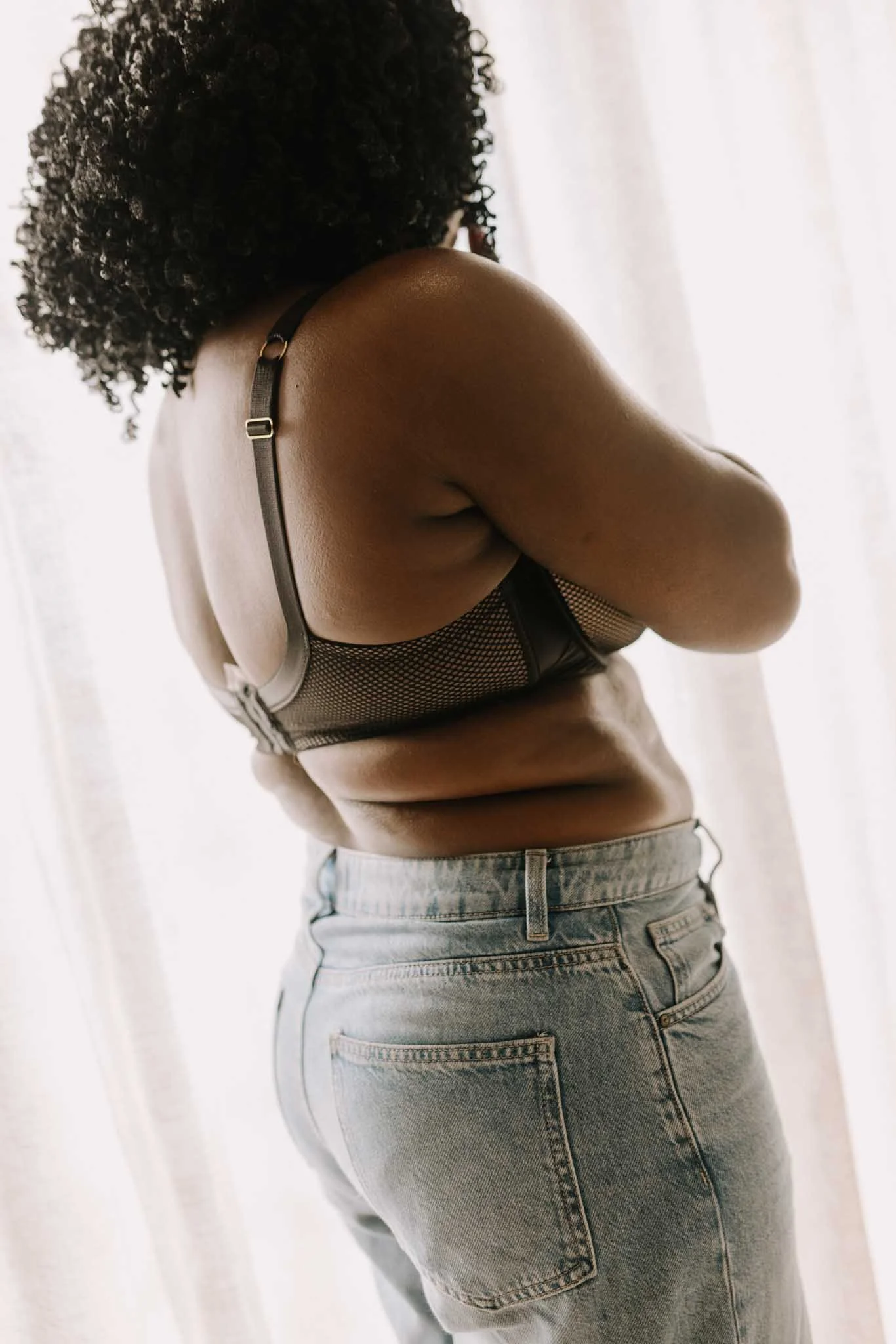 A person with curly black hair wearing a black mesh top and light blue jeans, standing near a white curtain.