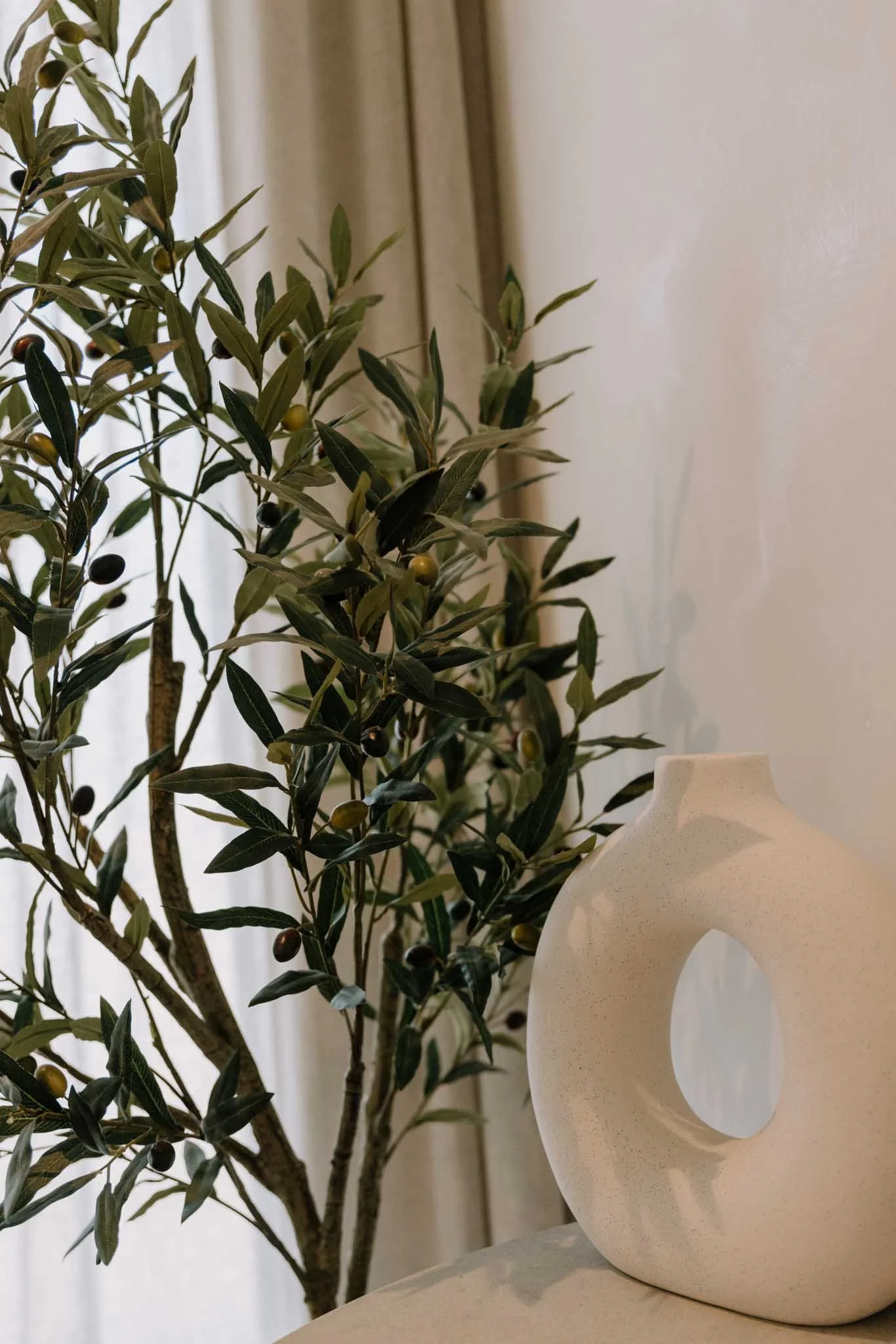 A decorative white ceramic vase with an unusual circular handle is placed next to a plant with green leaves and small black and green berries, set in front of a window with curtains.