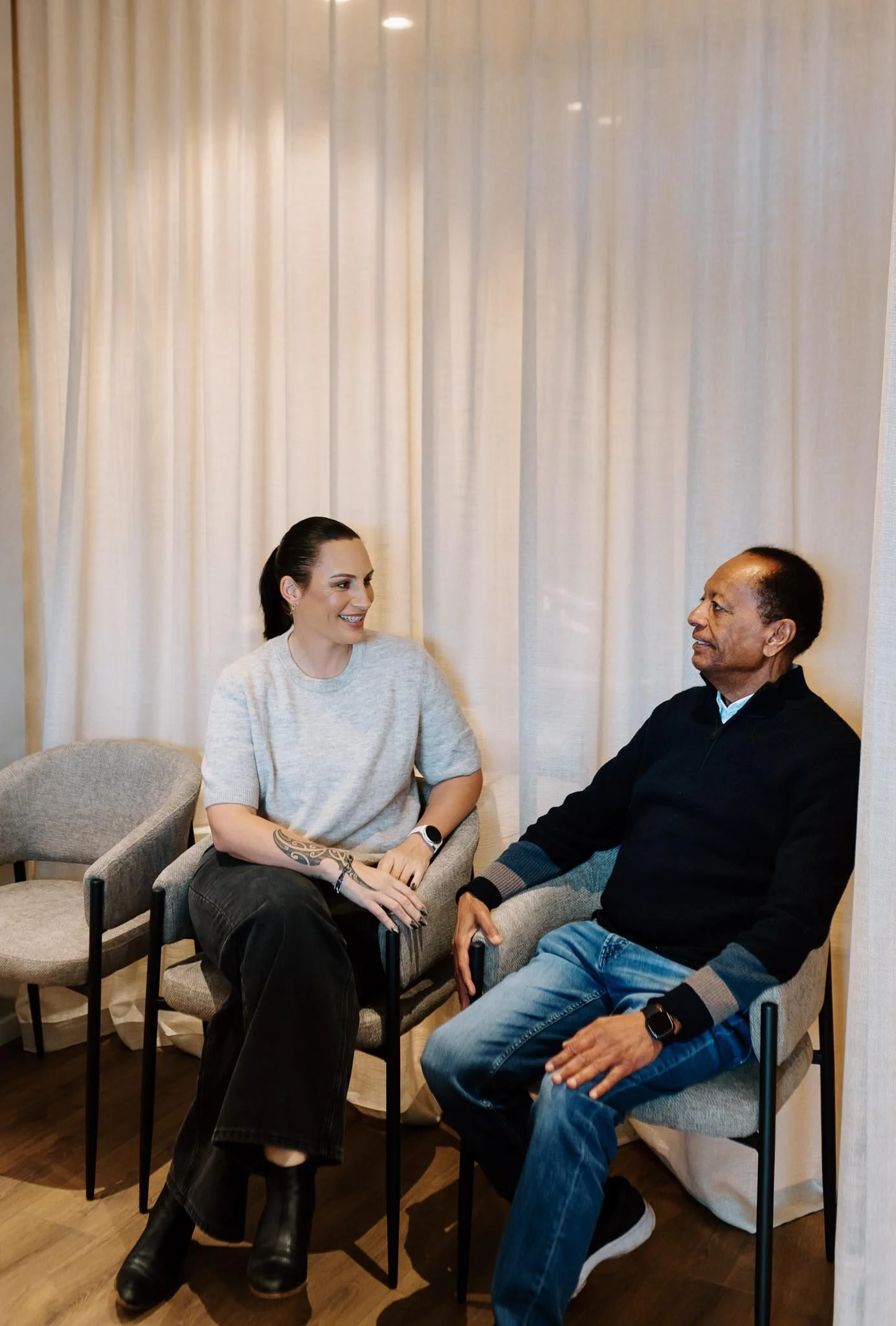 A woman and a man sit facing each other in conversation, inside a room with cream-colored curtains.