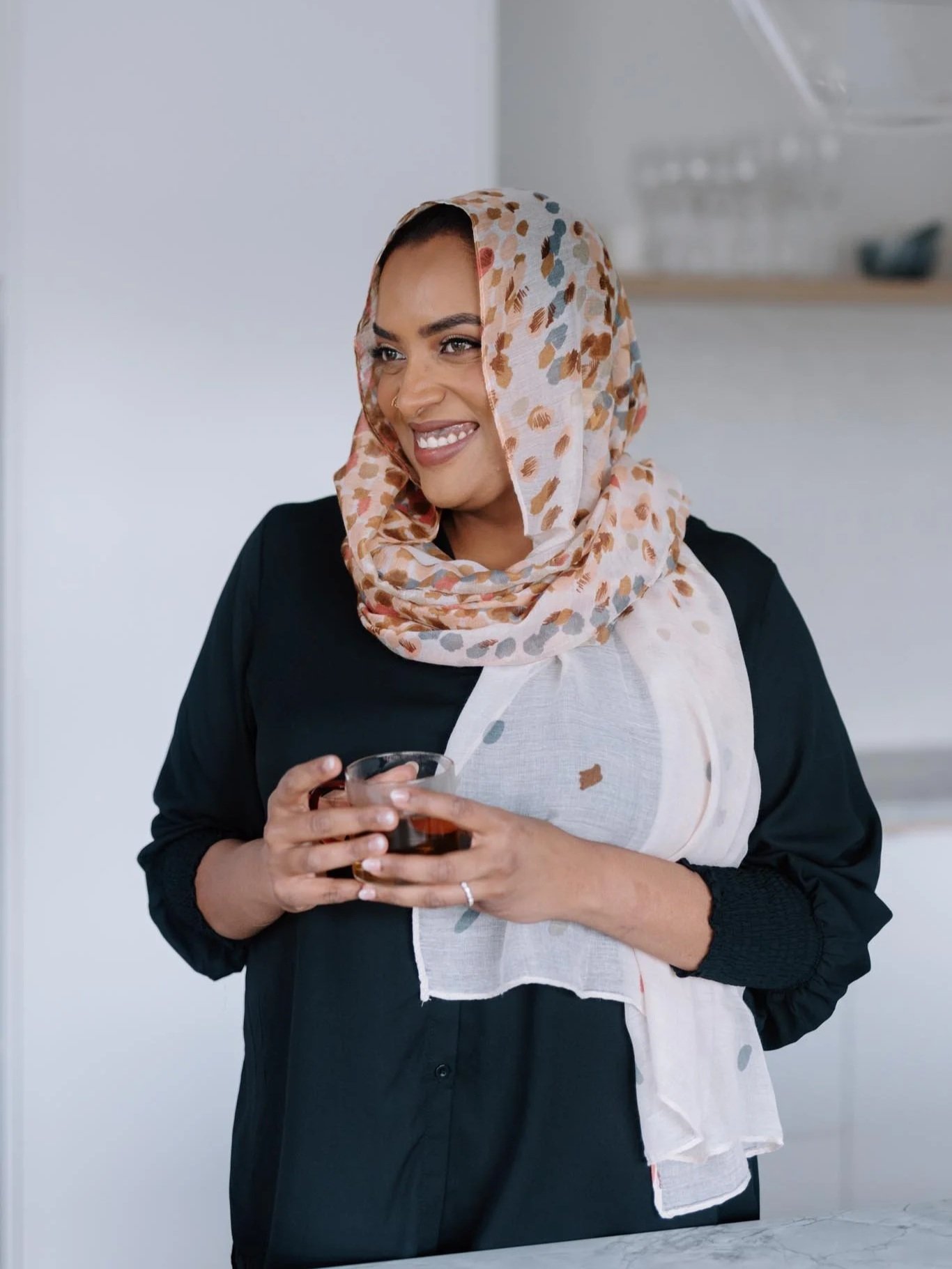 A woman with a cheerful expression, wearing a colorful headscarf, black jacket, and holding a glass of dark beverage in both hands, standing in a modern indoor setting.