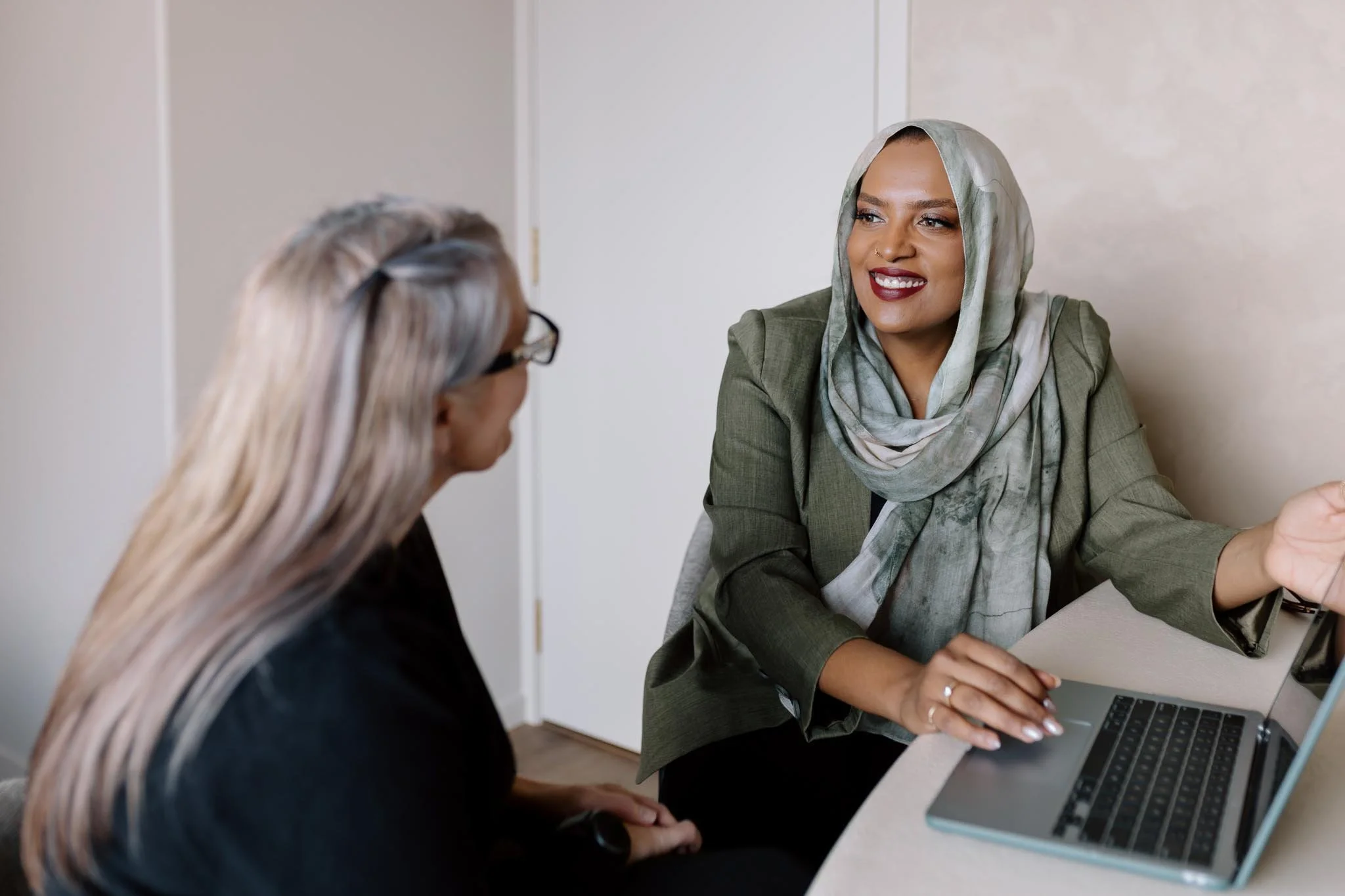 Two women having a conversation, one wearing a headscarf and working on a laptop, the other with gray hair, glasses, and a black top, sitting in a room with a beige wall.