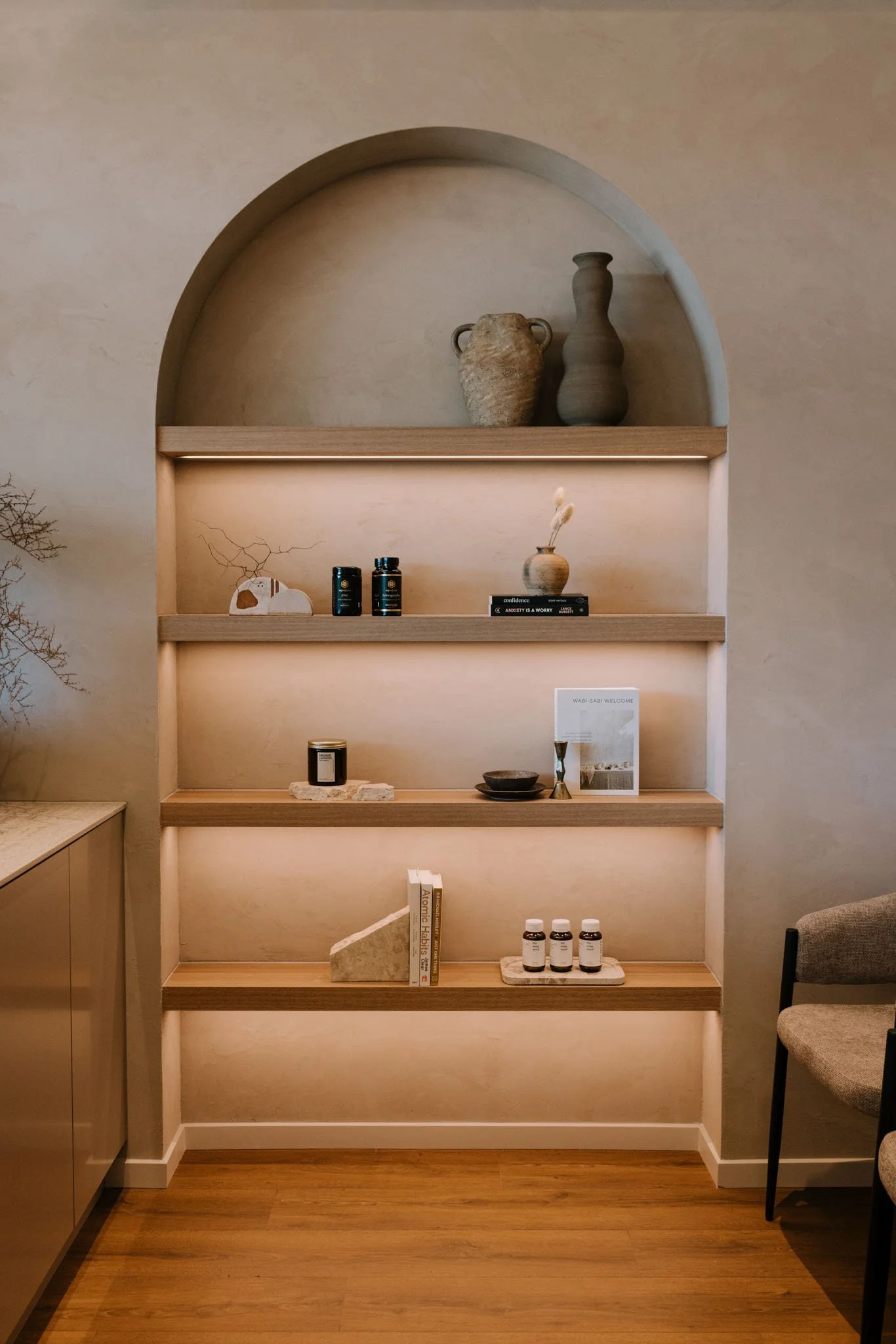 Empty wooden shelf with decorative items, books, and bottles in a room with wooden flooring and beige walls.