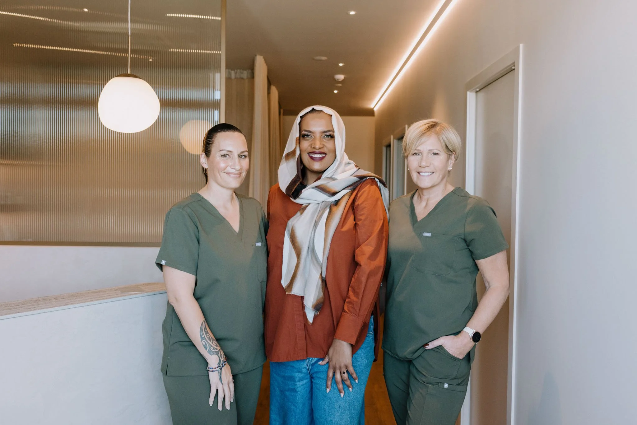 Three women standing together in a corridor. The woman in the center is wearing a headscarf and brown clothing, flanked by two women in green nurse scrubs. They are smiling at the camera.