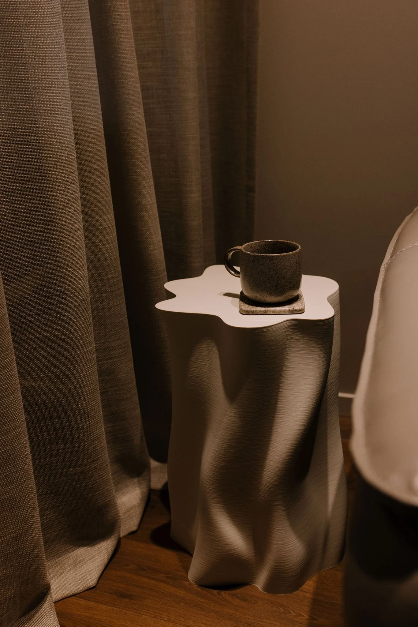 A small, round, white side table with a speckled gray mug on top is positioned next to a beige curtain and a white upholstered chair. The setting appears to be indoors with warm lighting.