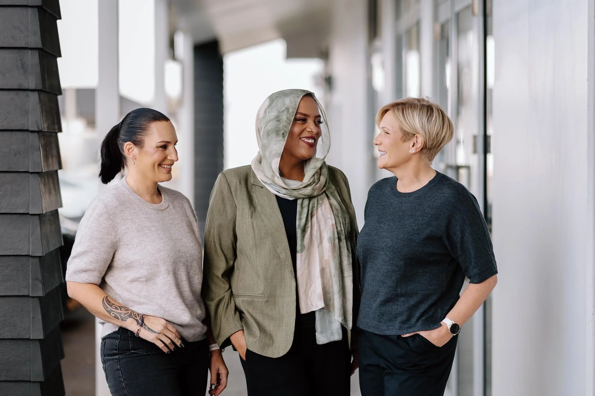 Three women are conversing outdoors near a modern building. They are smiling and appear to be enjoying their chat.