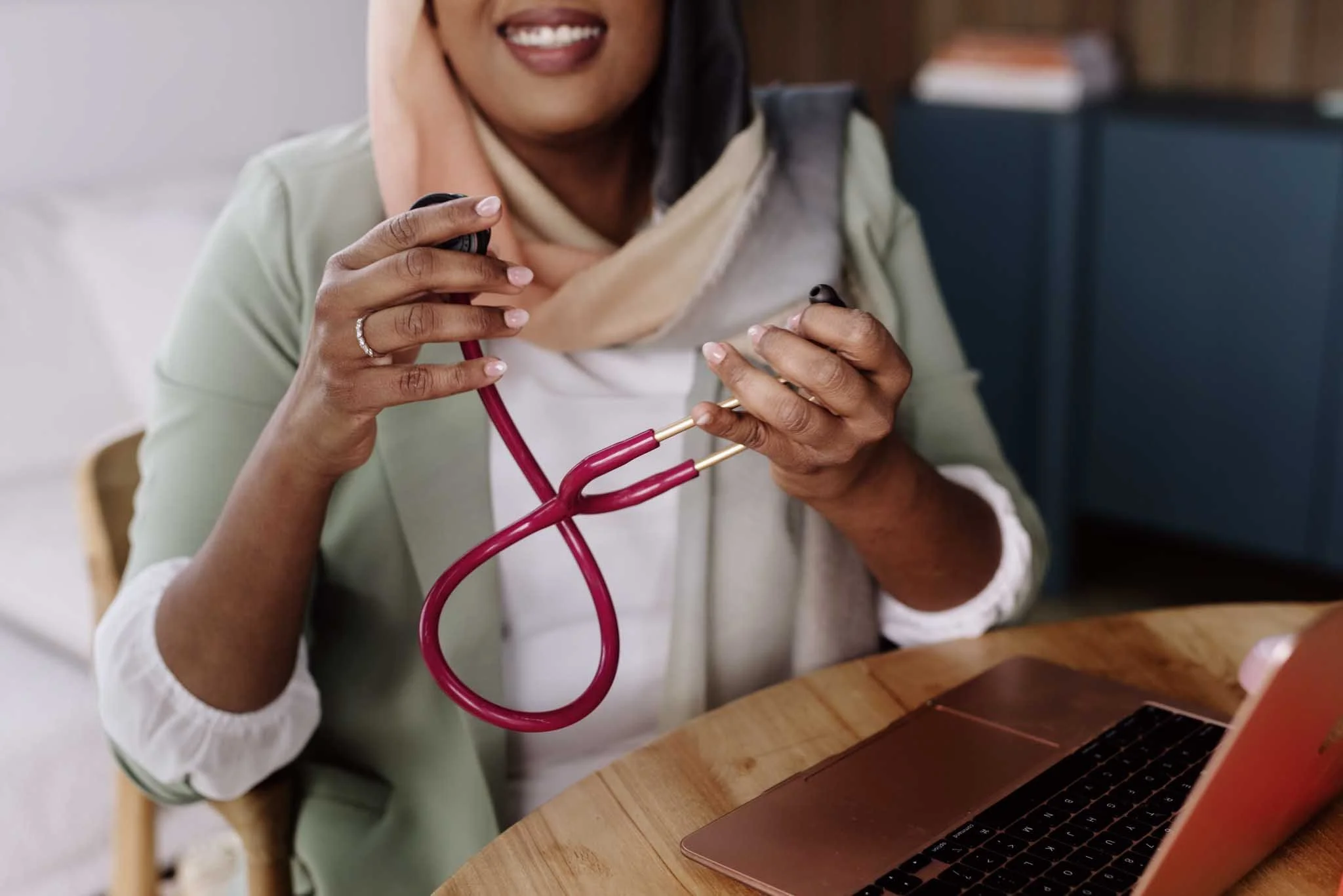 A woman smiling and holding a stethoscope near her ear while sitting at a wooden table with a laptop.
