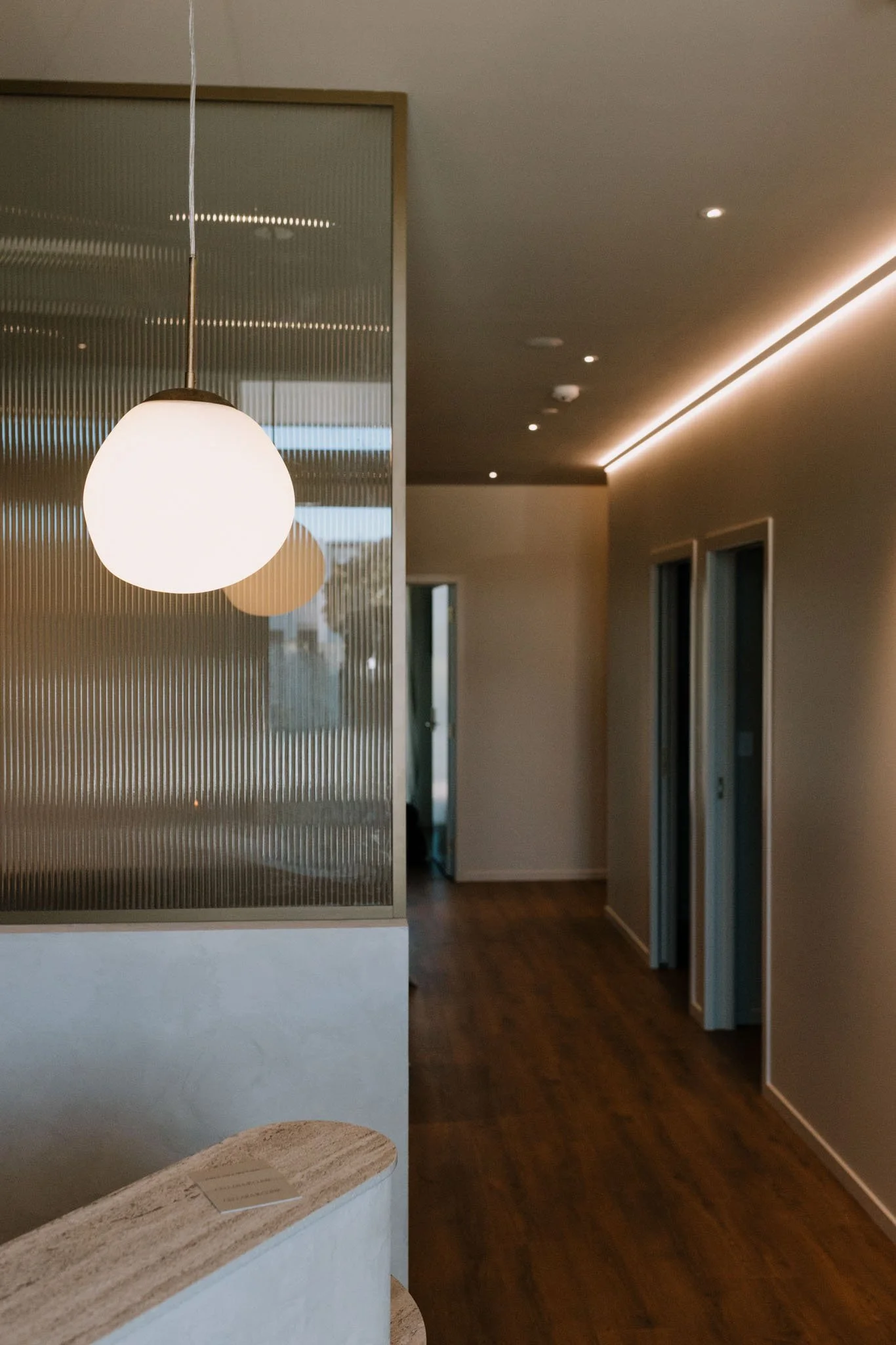Interior of a modern room with a hanging white spherical lamp, a wooden counter, and a textured glass partition. The room has wooden flooring, with small ceiling lights and LED strip lighting along the ceiling edge.