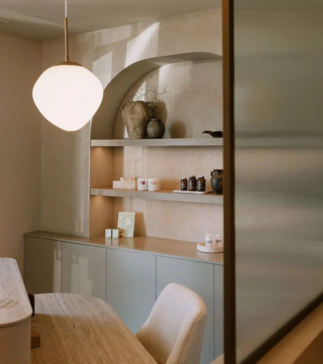 Interior view of a room with beige walls, displaying a built-in shelf with decorative vases, jars, and small objects, and a glass-fronted cabinet below. A soft glow of a hanging pendant light is visible, casting light on the shelf.