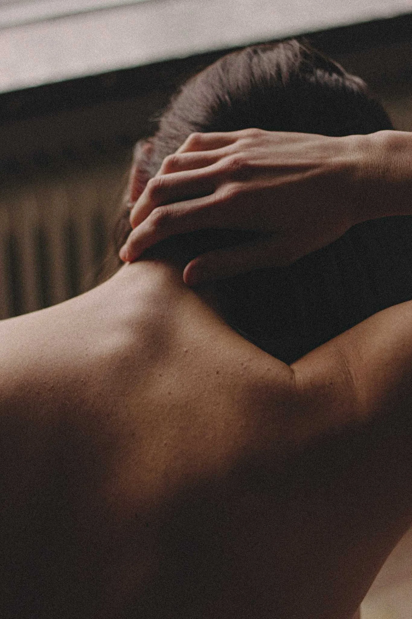 Close-up of a person with dark hair holding their head with one hand, resting on their shoulder in a dark setting.