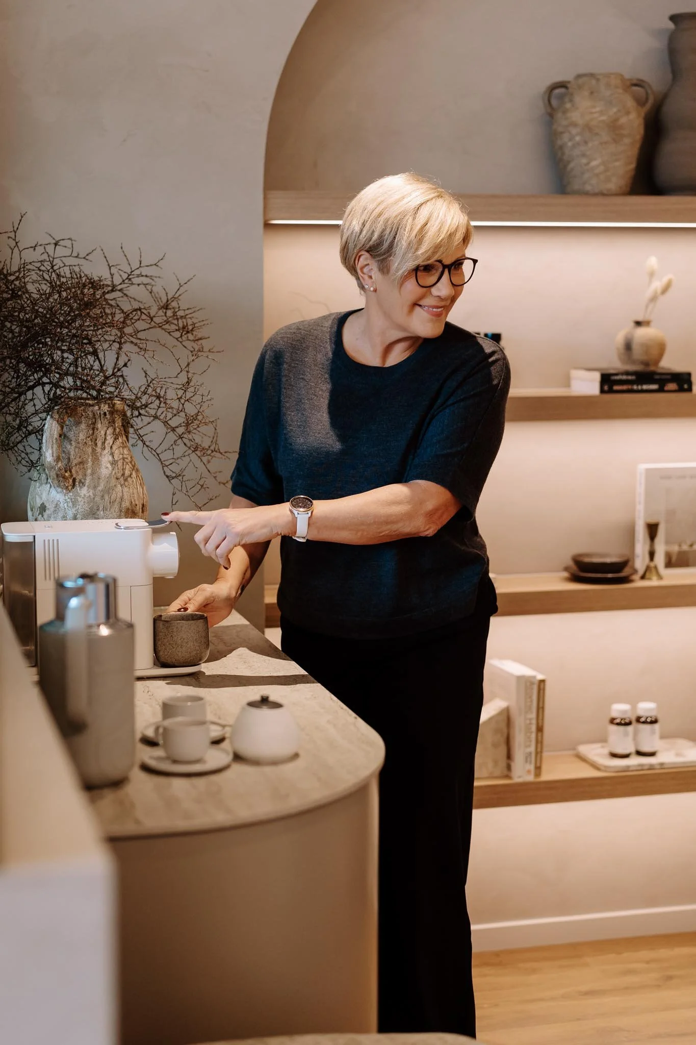 A woman with short gray hair and glasses smiling while standing in a kitchen near a coffee machine, with decorative vases and shelves in the background.