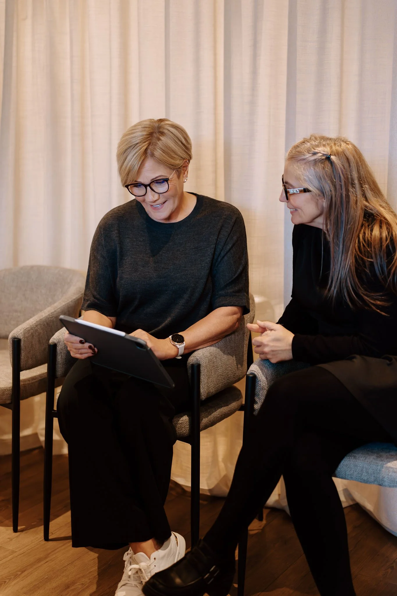 Two women sitting and looking at a tablet together in a room with beige curtains. One woman has short blond hair and is wearing glasses, a black top, and white sneakers. The other woman has long blond hair, glasses, and is wearing a black top and black pants.